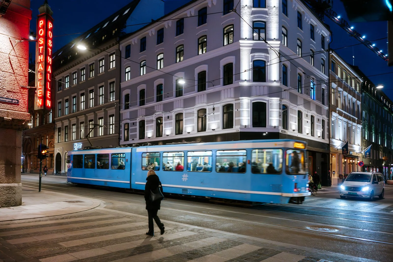 Oslo at night: a dynamic interplay of urban light. A blue tram glides past a crisp white facade, its motion marked by light streaks. The iconic 'Posthallen' neon casts a warm, historic glow against the cool Nordic nocturnal charm.