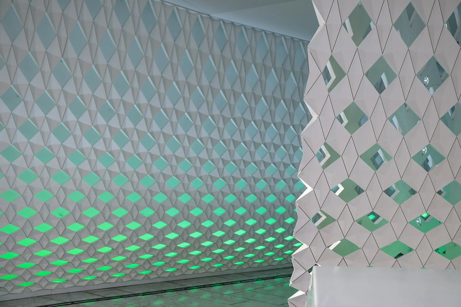 Futuristic white wall with geometric diamond-shaped cutouts, illuminated with green lights, and a white decorative partition with a similar pattern, partially blocking the view.