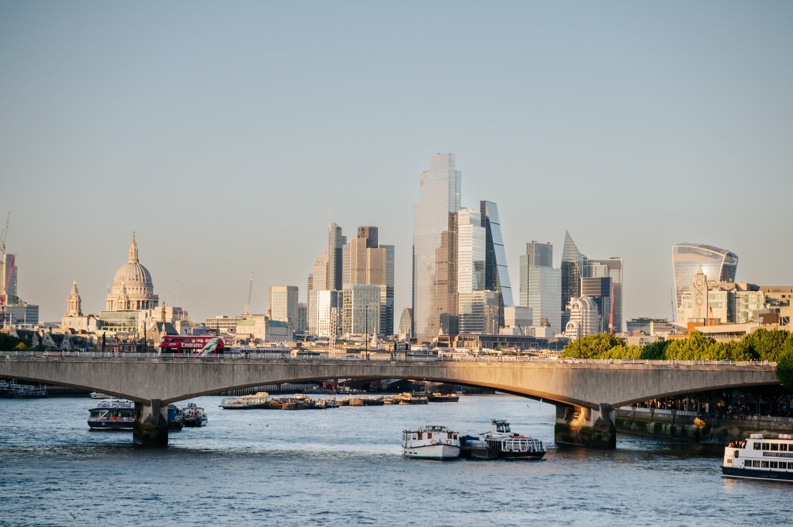 View of London skyline with buildings and the river Thames in the foreground, including boats and a bridge.