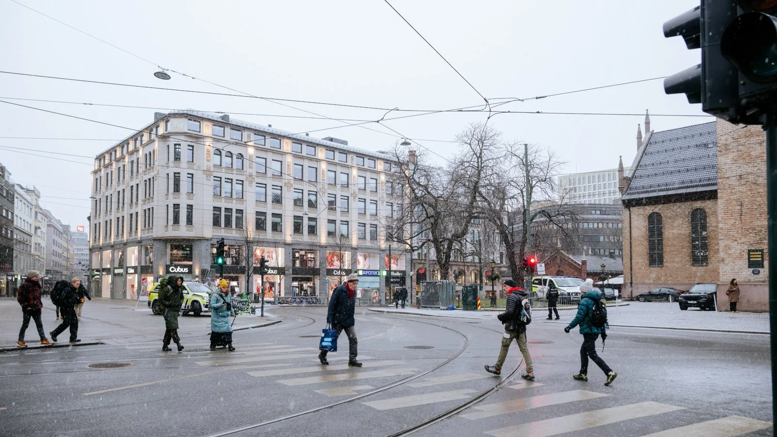People crossing a city street on a rainy day with buildings, traffic lights, and tram wires overhead.