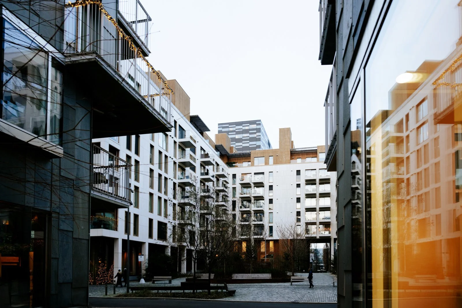 View of a modern residential courtyard surrounded by high-rise buildings with white and gray exteriors, balconies, and large glass windows. There are benches, trees, and a few people walking through the area, with reflections visible on the glass sur