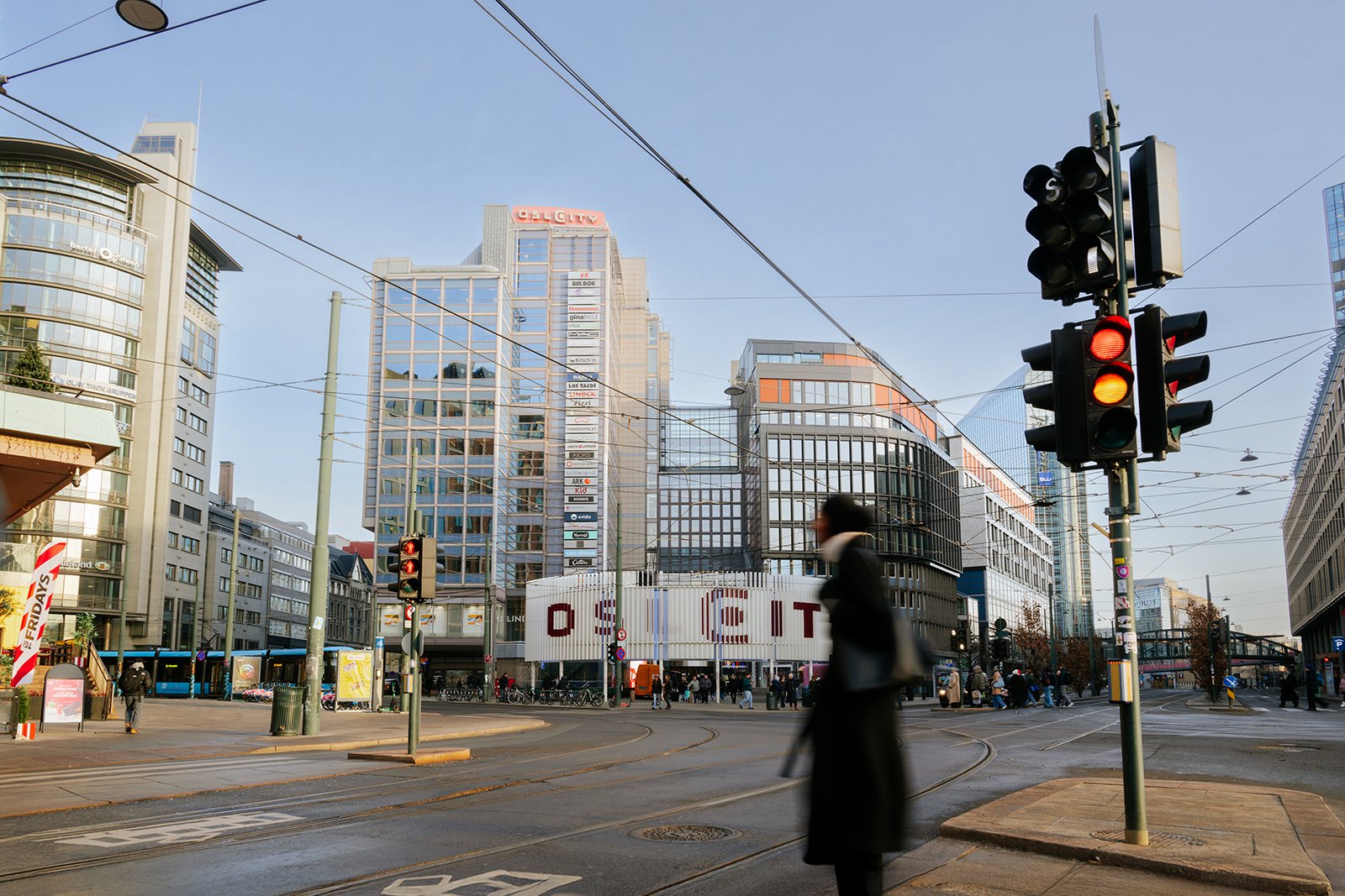 Urban city street scene with tall modern buildings, traffic lights showing red, tram tracks, pedestrians, and a woman walking with a bag.