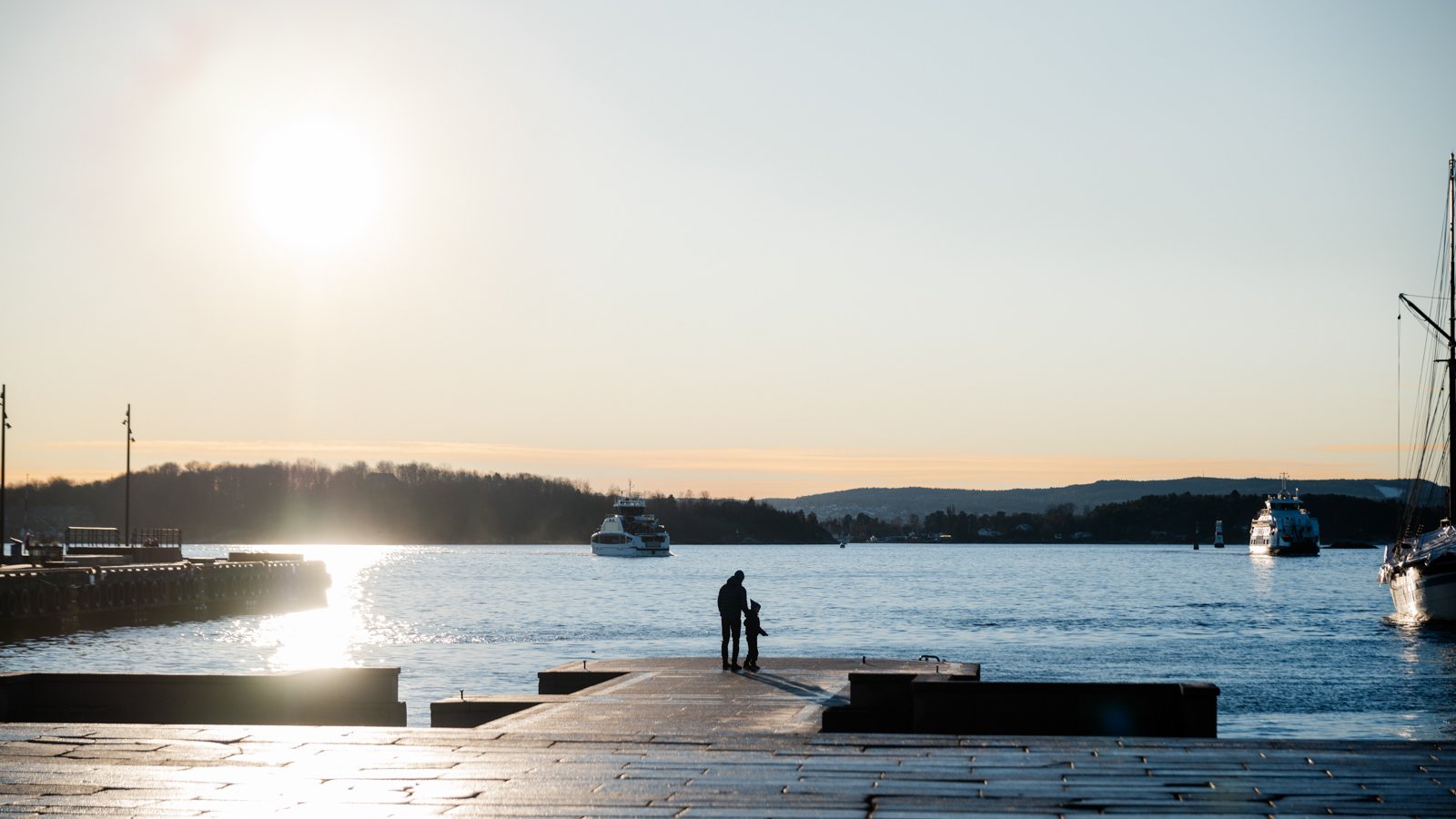 A silhouette of an adult and a child walking on a dock near a body of water, with boats sailing in the distance and the sun setting or rising, creating a reflective glow on the water surface.