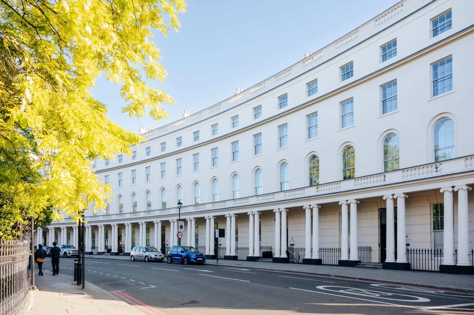 Street view of a large white building with multiple windows, classical columns, and iron fencing, with a few parked cars and two pedestrians walking.
