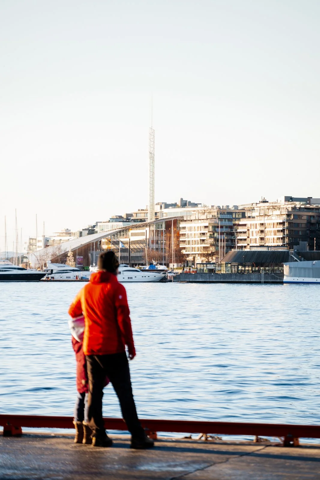A father and son share a quiet moment on the Oslo waterfront, their figures serving as a warm splash of color against the cool Nordic light. They gaze towards the modern urban skyline and the bustling harbor, where yachts and new architecture converg