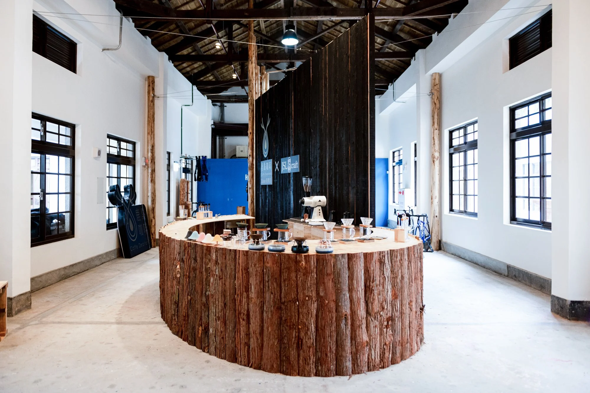 Interior of a coffee shop with a round wooden counter made from logs, display of coffee cups and brewing equipment, white walls, large windows, and a black divider with signs on it.