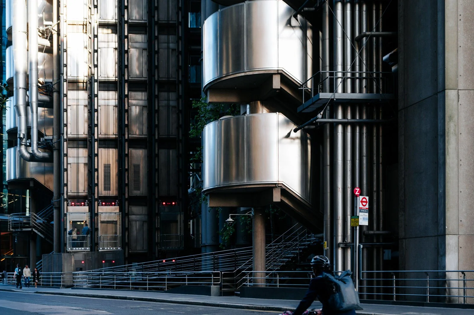 Modern urban building with metallic exterior and external staircase, surrounded by street with pedestrians and a cyclist.
