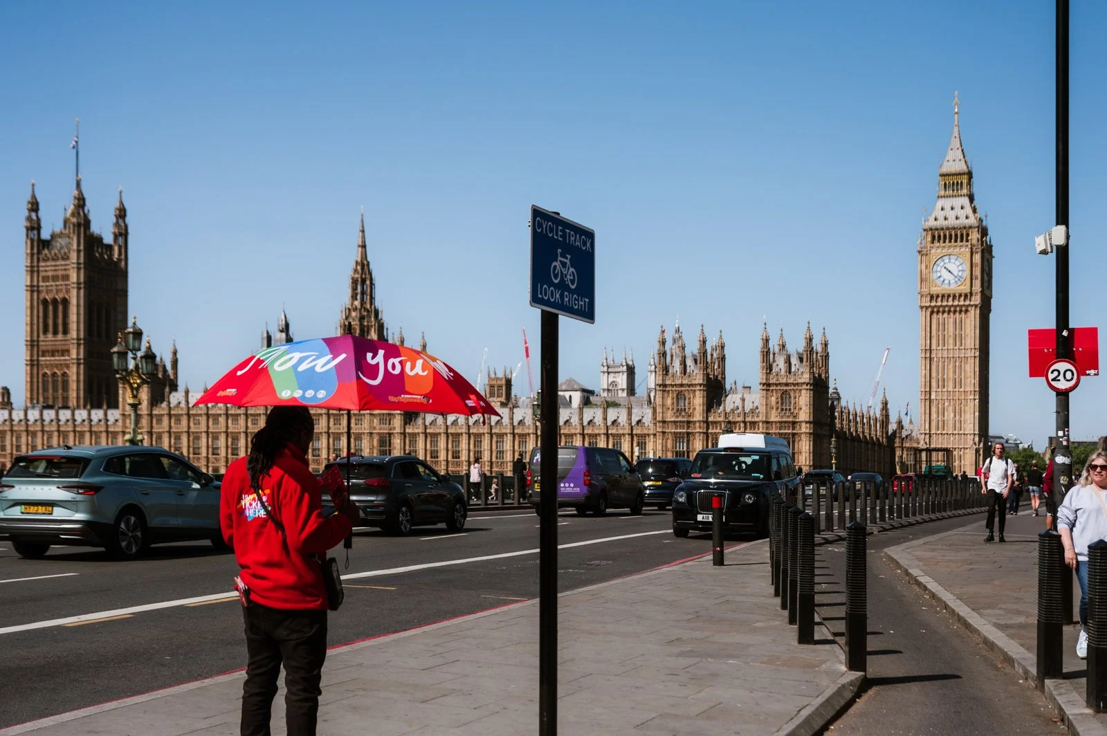 A person holding a colorful umbrella with the words "How You" on it, walking on the sidewalk near Westminster Palace and Big Ben in London, with cars on the street and blue sky overhead.