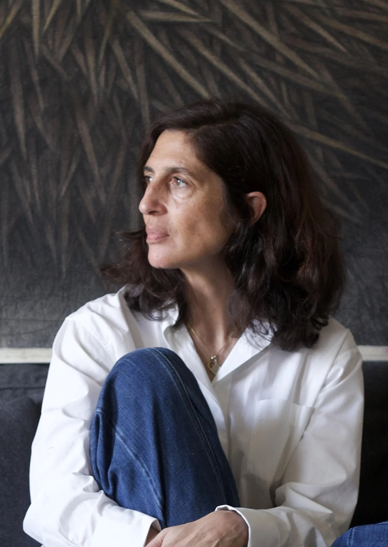 A woman with dark wavy hair wearing a white shirt and blue jeans sitting against a dark textured wall, looking to her left.