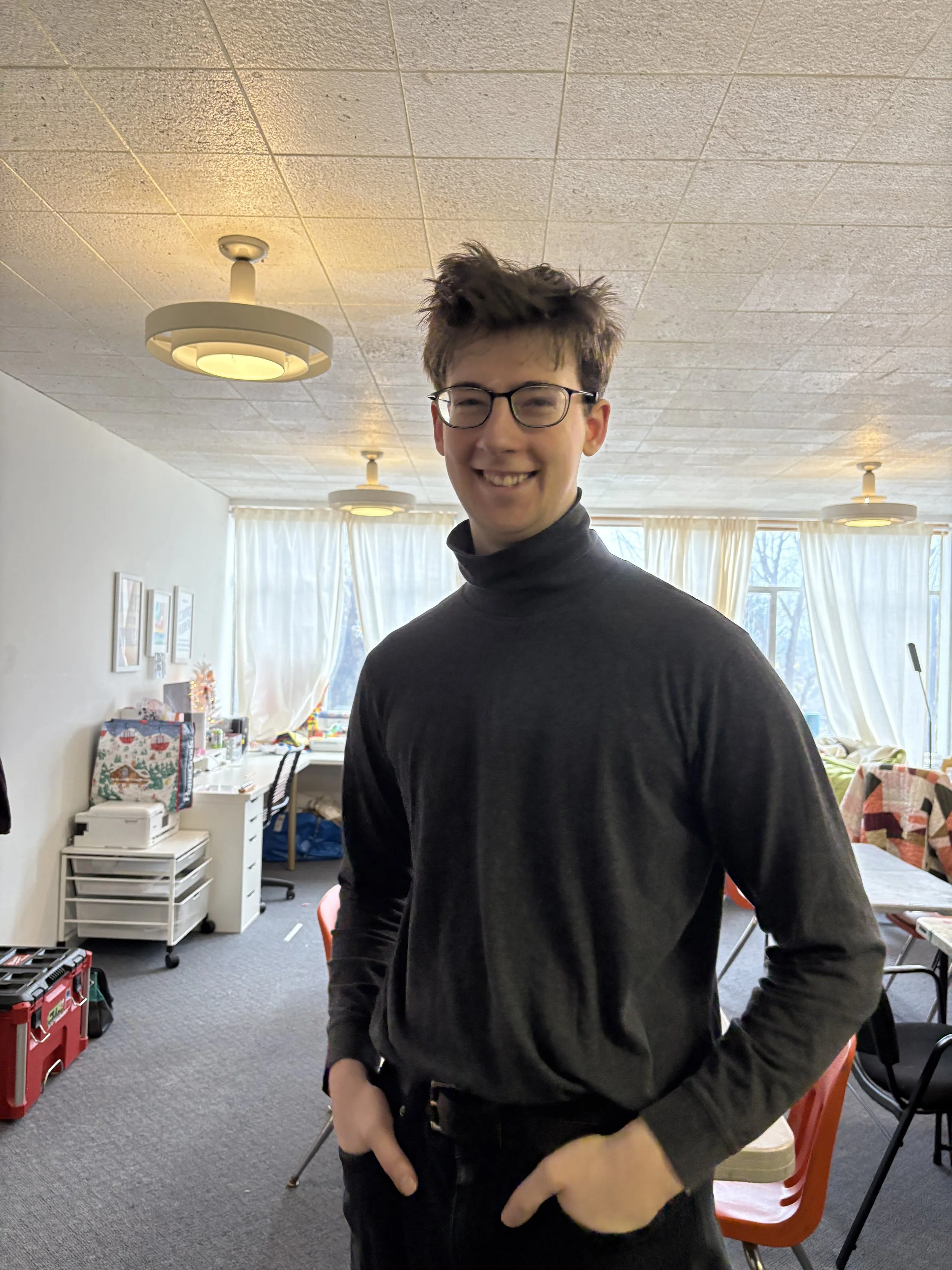 A young man with glasses and messy hair smiling, standing in a room with large windows, curtains, and tables.