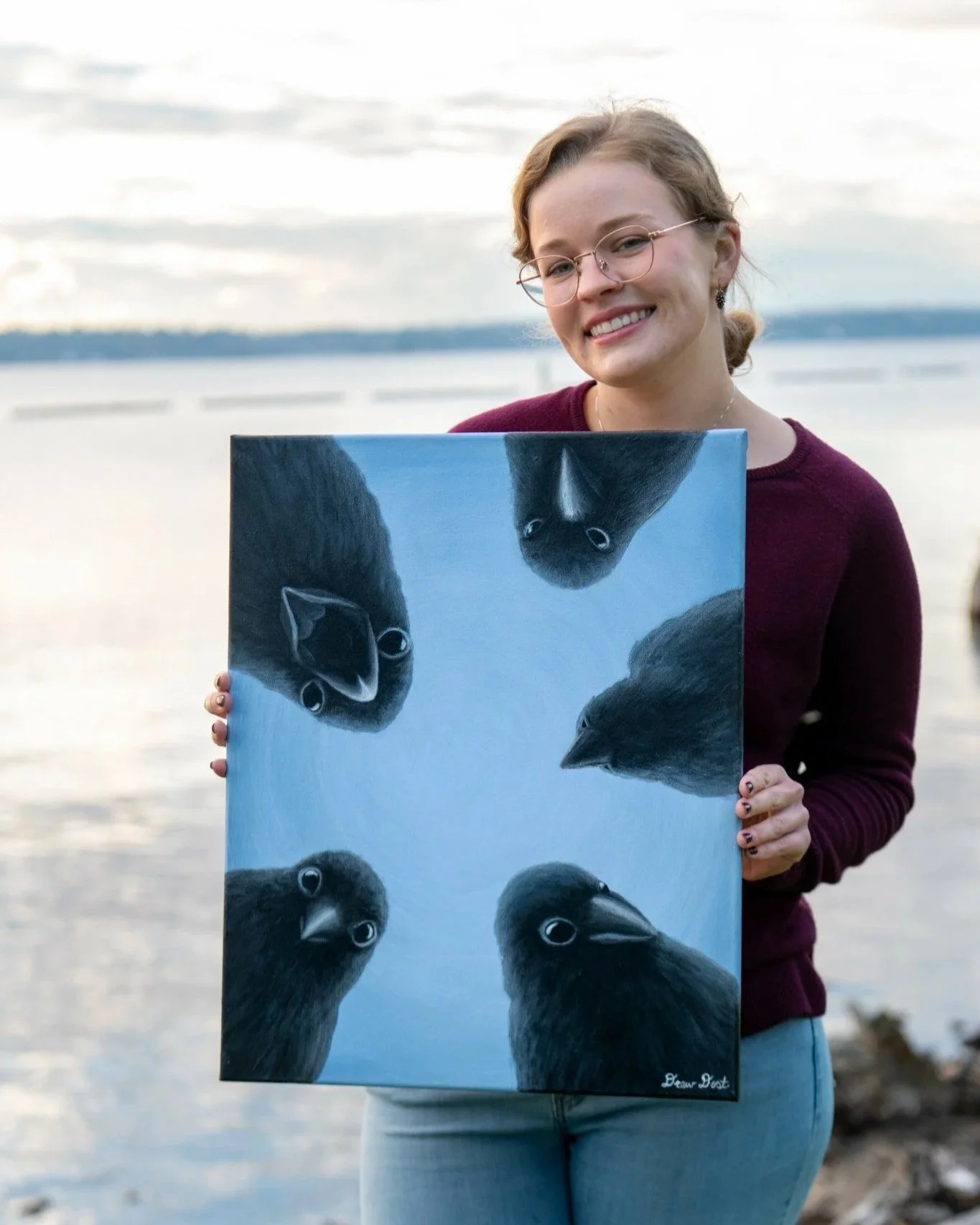 Drew smiling at the camera while holding her painting, Life of a Coin, featuring five crows looking down at the viewer in a circle.