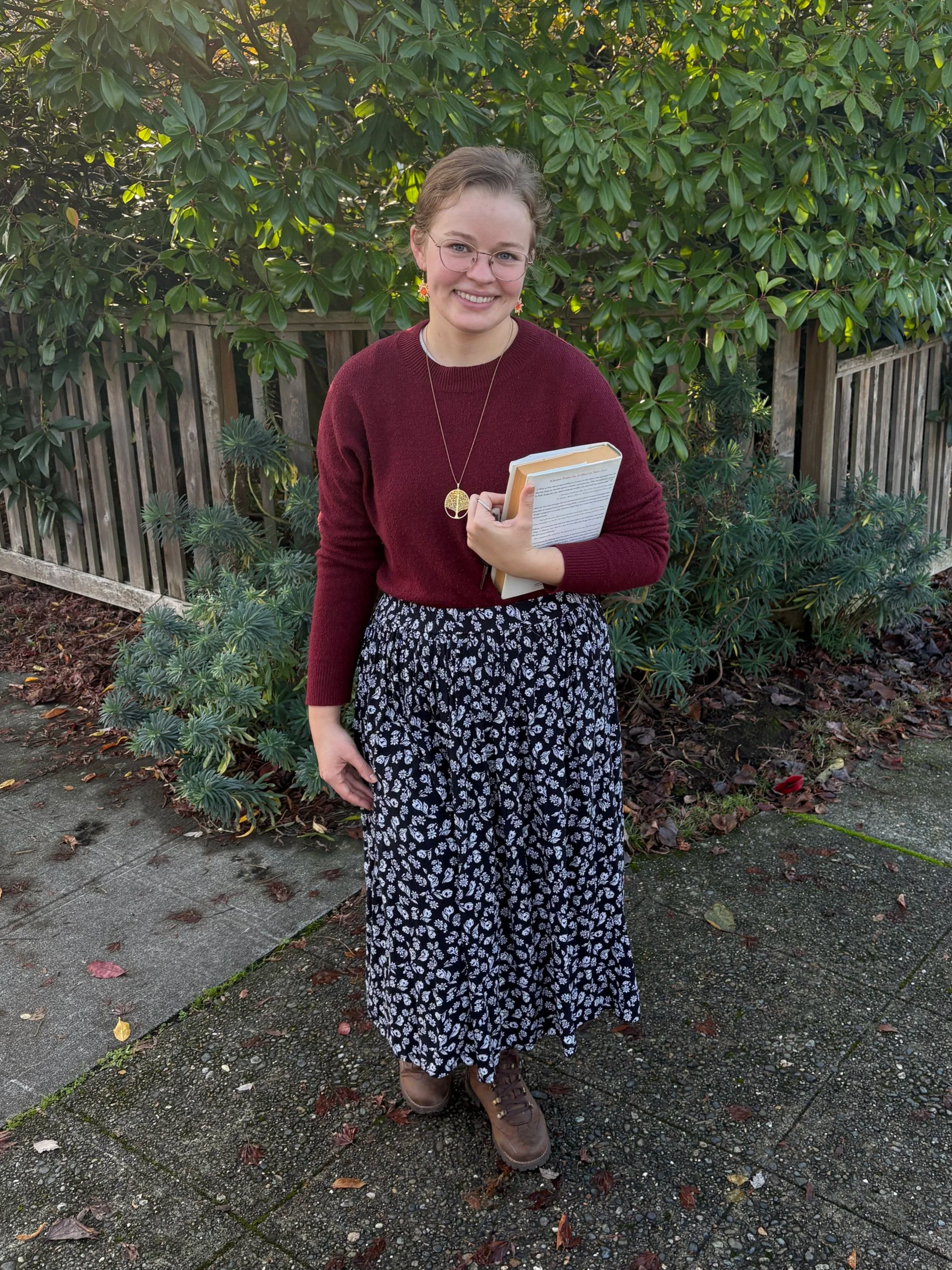 A young woman with glasses, holding books, standing outdoors on a concrete sidewalk with fallen leaves, in front of green bushes and a wooden fence, smiling at the camera.