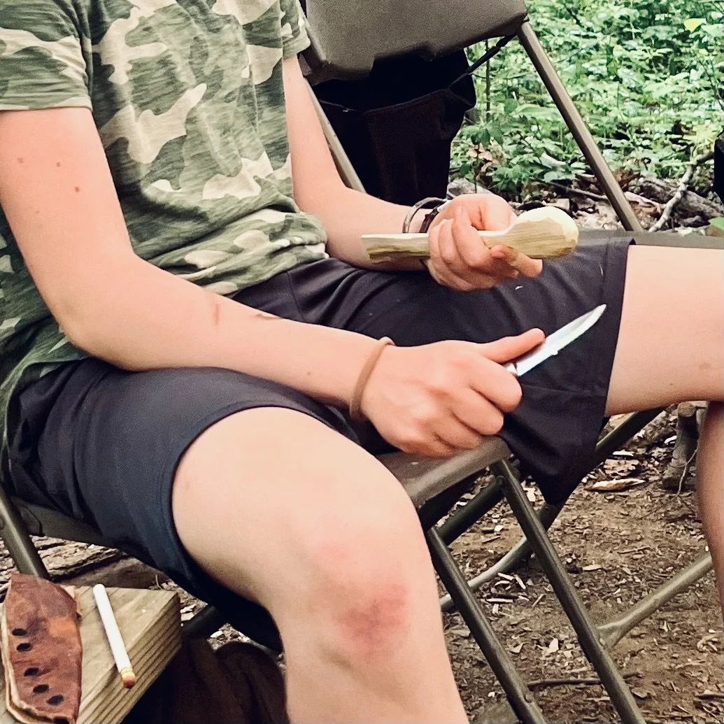 A teen carves a wooden spoon with a knife at a screen-free outdoor summer camp for teens near Asheville, NC.