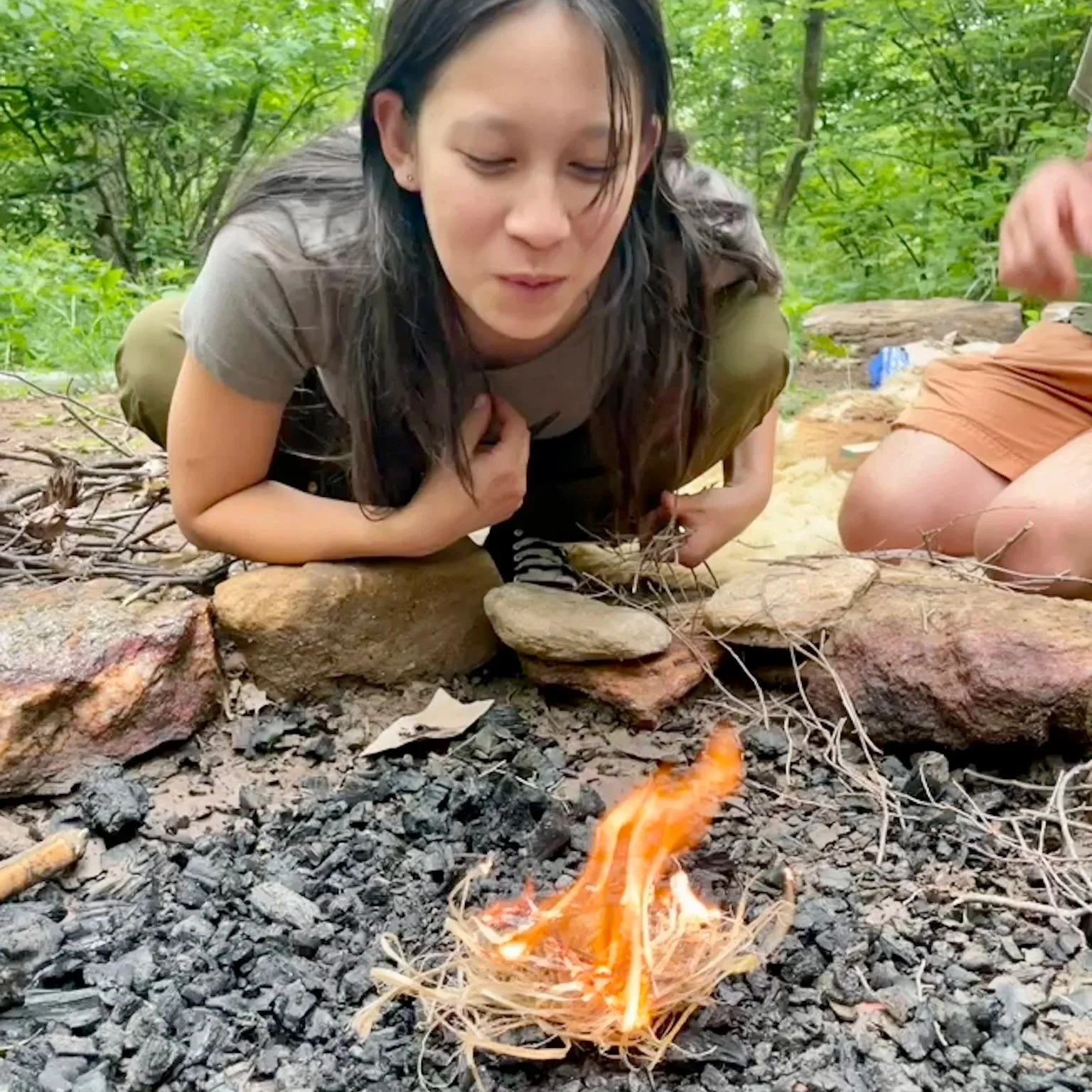 A teen girl smiles as she blows a flame to life after the group made a fire by friction at a screen-free outdoor camp near Asheville, NC.