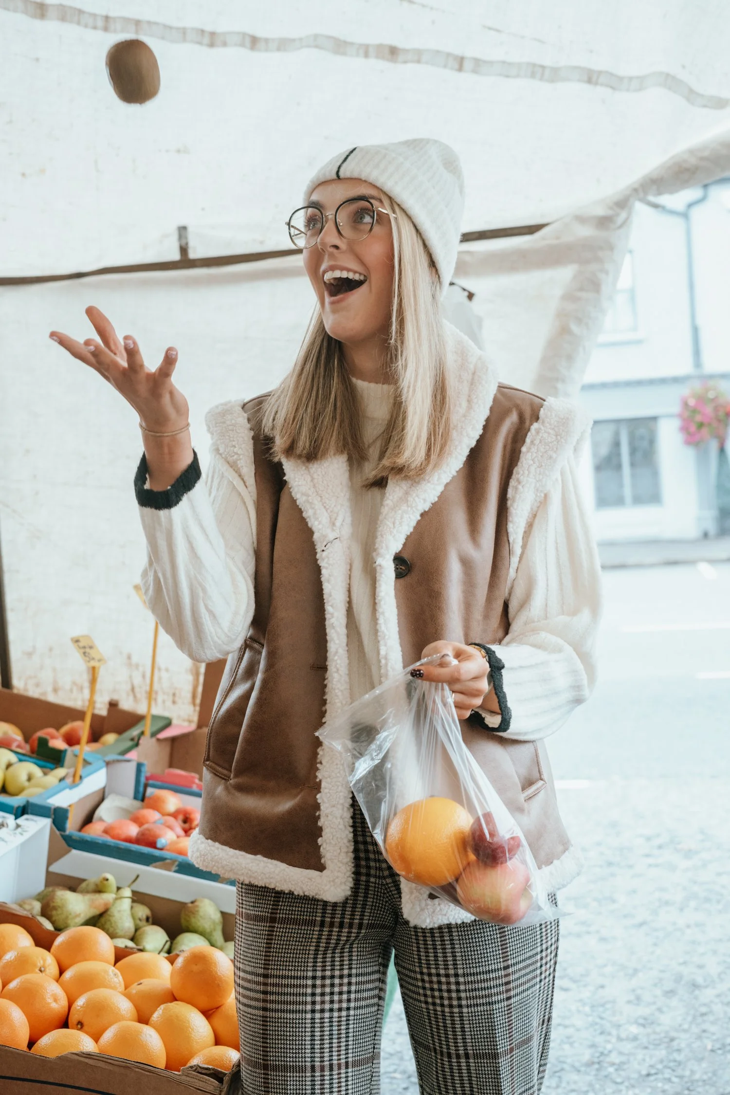 A young woman at an outdoor fruit market, tossing an apple into the air while holding a plastic bag with various fruits.