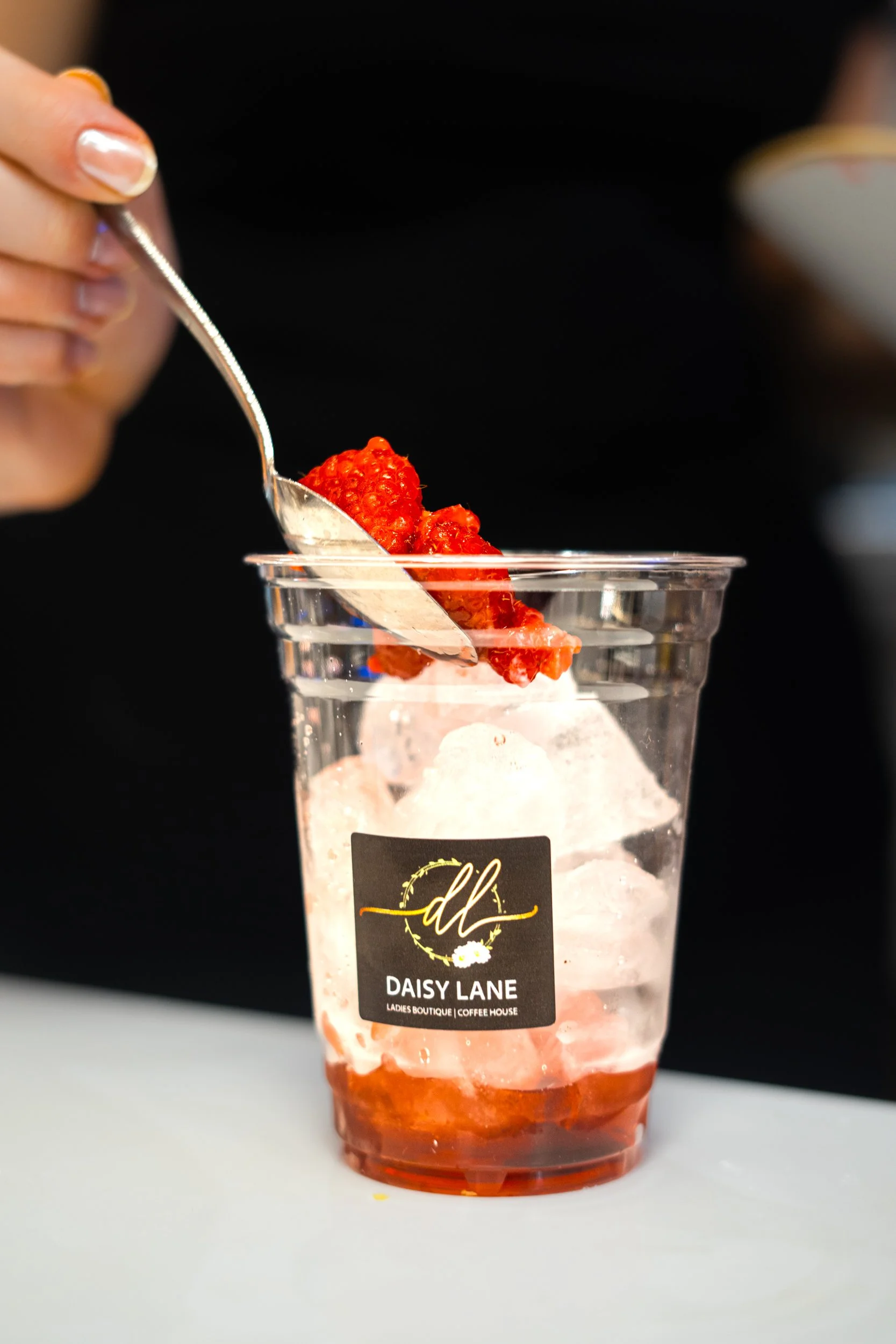 A hand with painted nails holding a spoon above a clear plastic cup filled with ice, strawberries, and a red syrup, with a "Daisy Lane" logo on the cup.