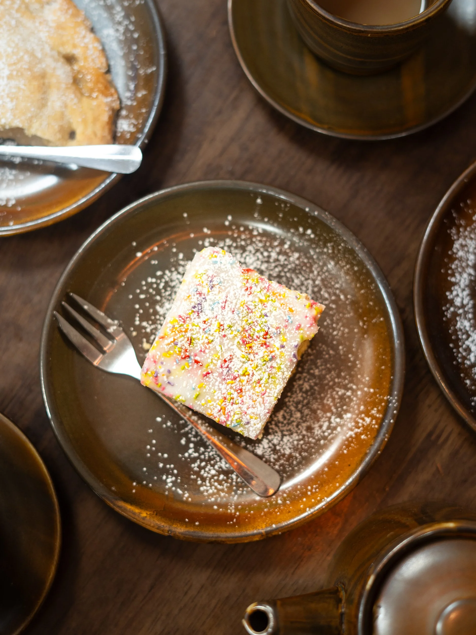 A square piece of birthday cake with white icing and colorful sprinkles on a small dark plate, with a fork resting on the side of the cake and sprinkled powdered sugar or decorative powder around it.
