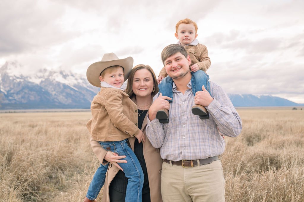 Family, mom, dad, two sons, mom holding the one with a cowboy hat and the other on dads shoulder, smiling while on vacation at Jackson Hole