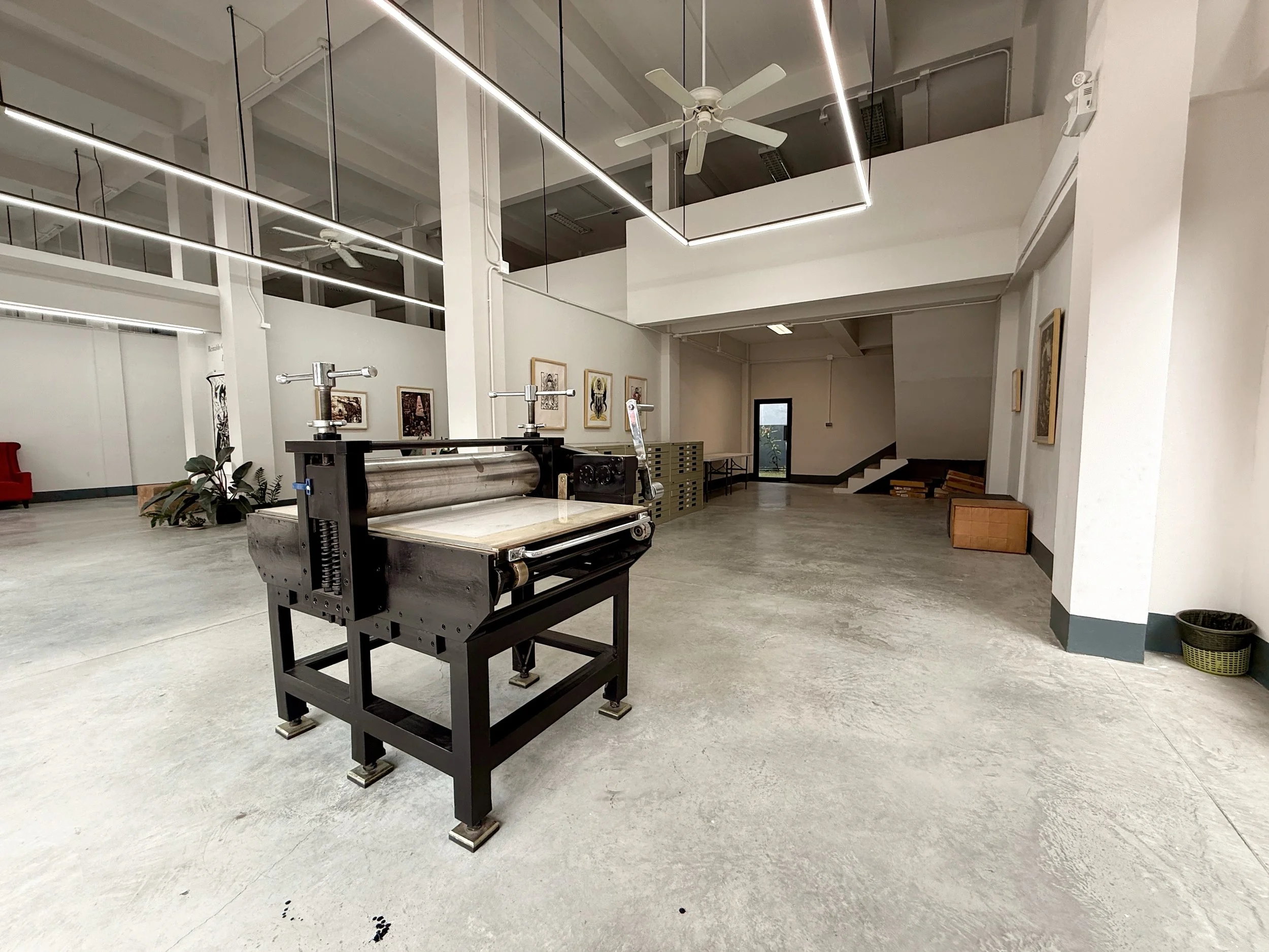 Interior of an art gallery or museum with white walls, framed artworks, and bright linear ceiling lights. A small machine or printing press is in the foreground, with a potted plant next to it. There are stairs and a door in the background.