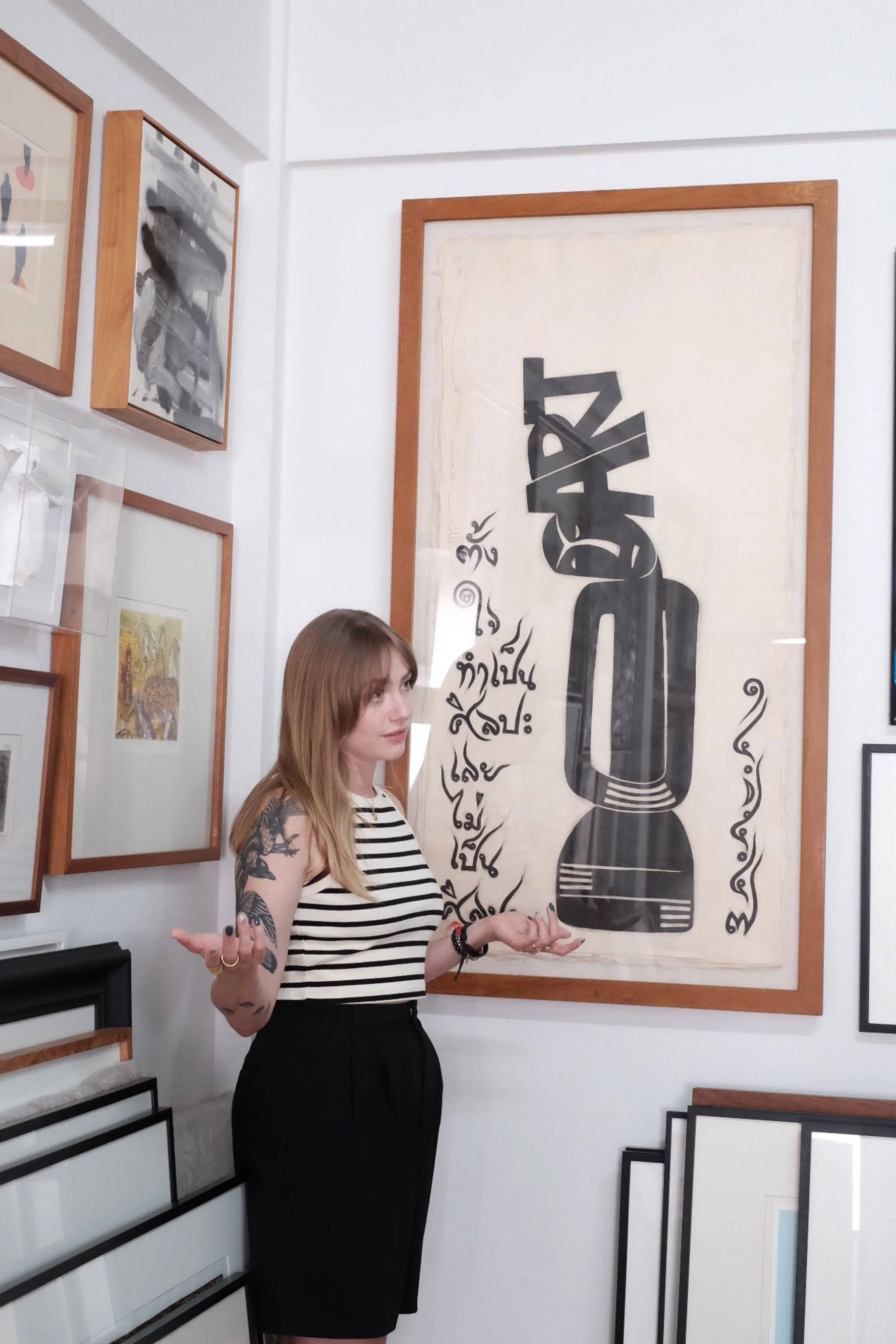 A woman with long blonde hair and extensive black tattoos on her arms is standing in an art gallery, gesturing as if explaining something. She is wearing a black and white striped sleeveless top and black pants. Behind her, there is a large piece of artwork with black abstract figures and various swirling lines on a light-colored background, framed in wood.