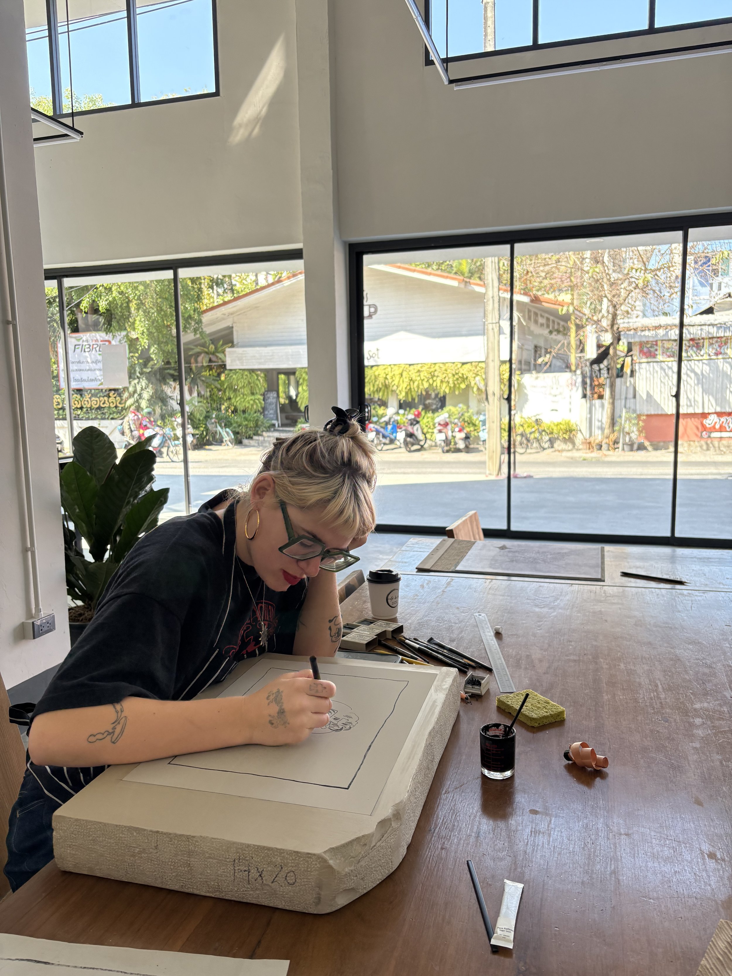 A woman with short blonde hair, wearing glasses and a black shirt, is sitting at a wooden table drawing on a large foam piece with a black marker. The foam has some markings and writing on it. The table has various art supplies, including brushes, a ruler, and a cup of black ink. There is a large window in the background showing a street scene with bicycles, trees, and buildings outside.