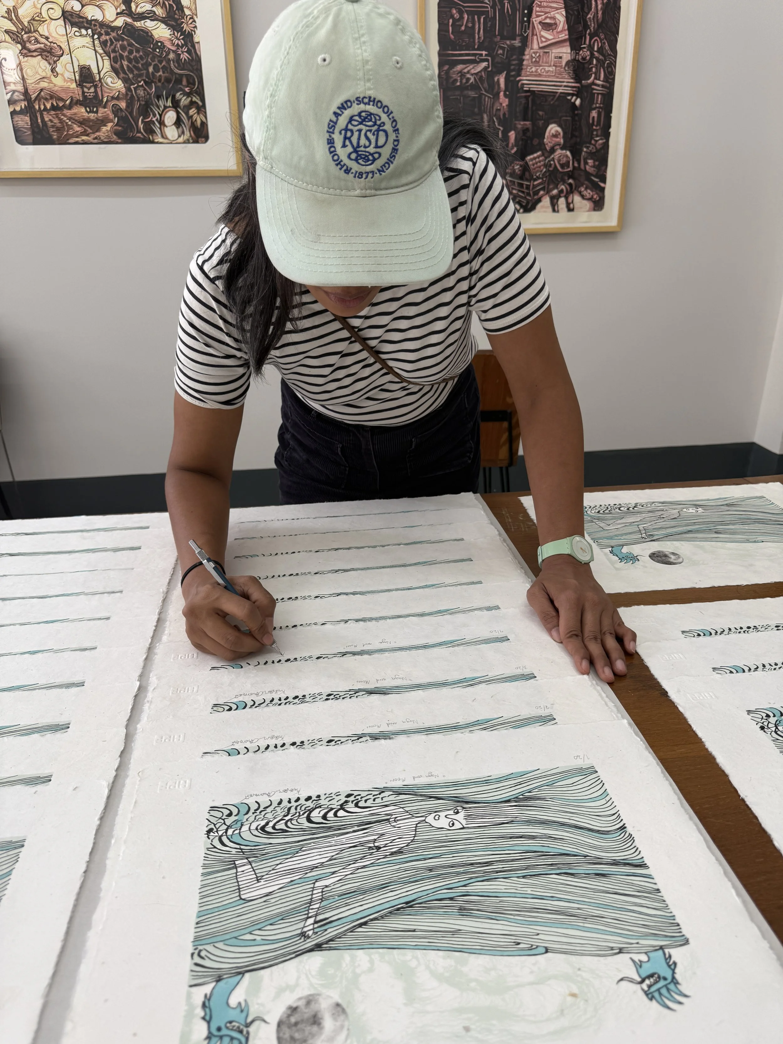 A person wearing a striped shirt and a white cap is signing a large, illustrated map or poster on a table. The background shows framed artwork on a wall.