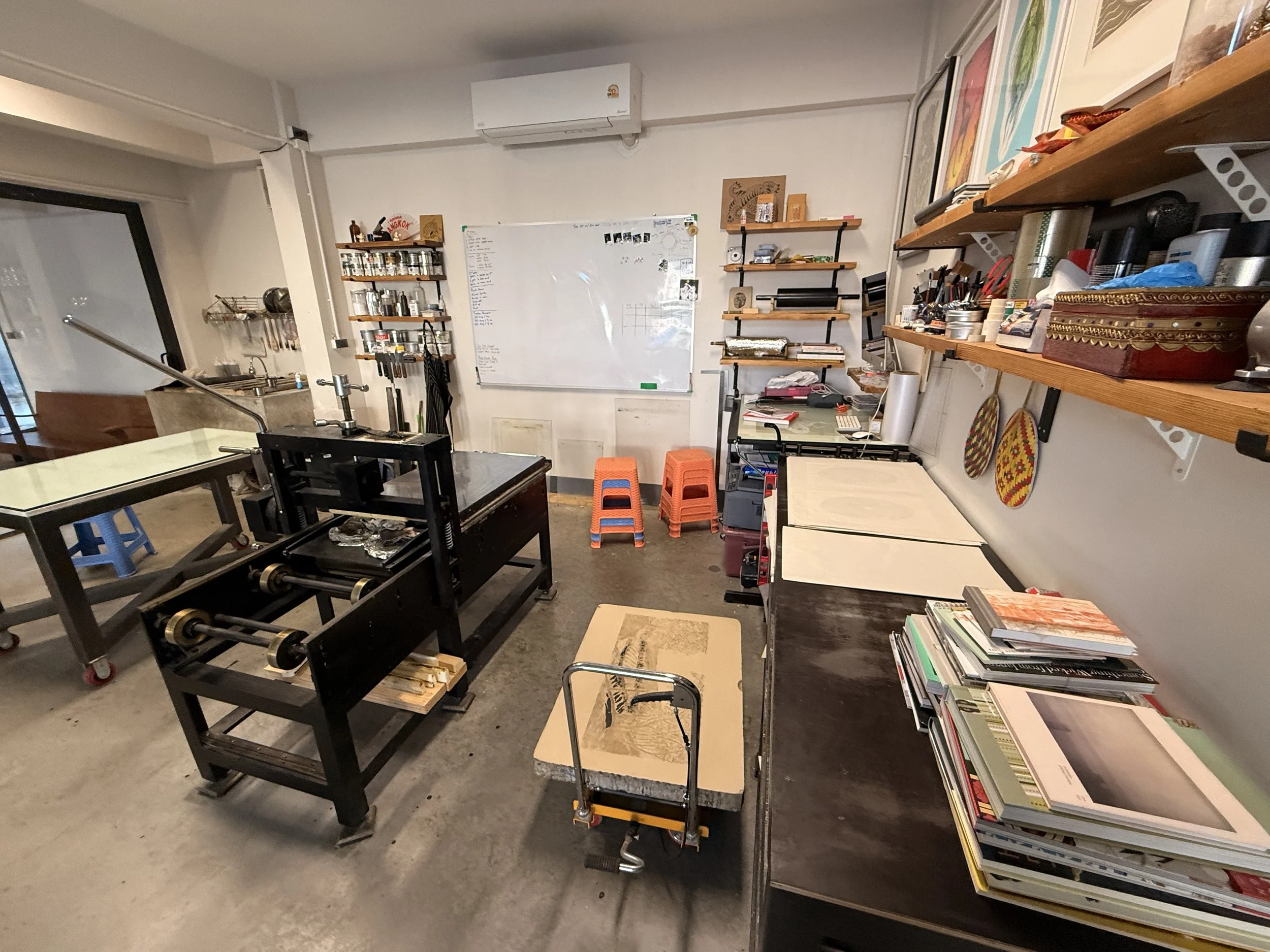 A cluttered creative workspace with shelves of art supplies, a whiteboard, books, and a foosball table, in a room with beige concrete floors and white walls.
