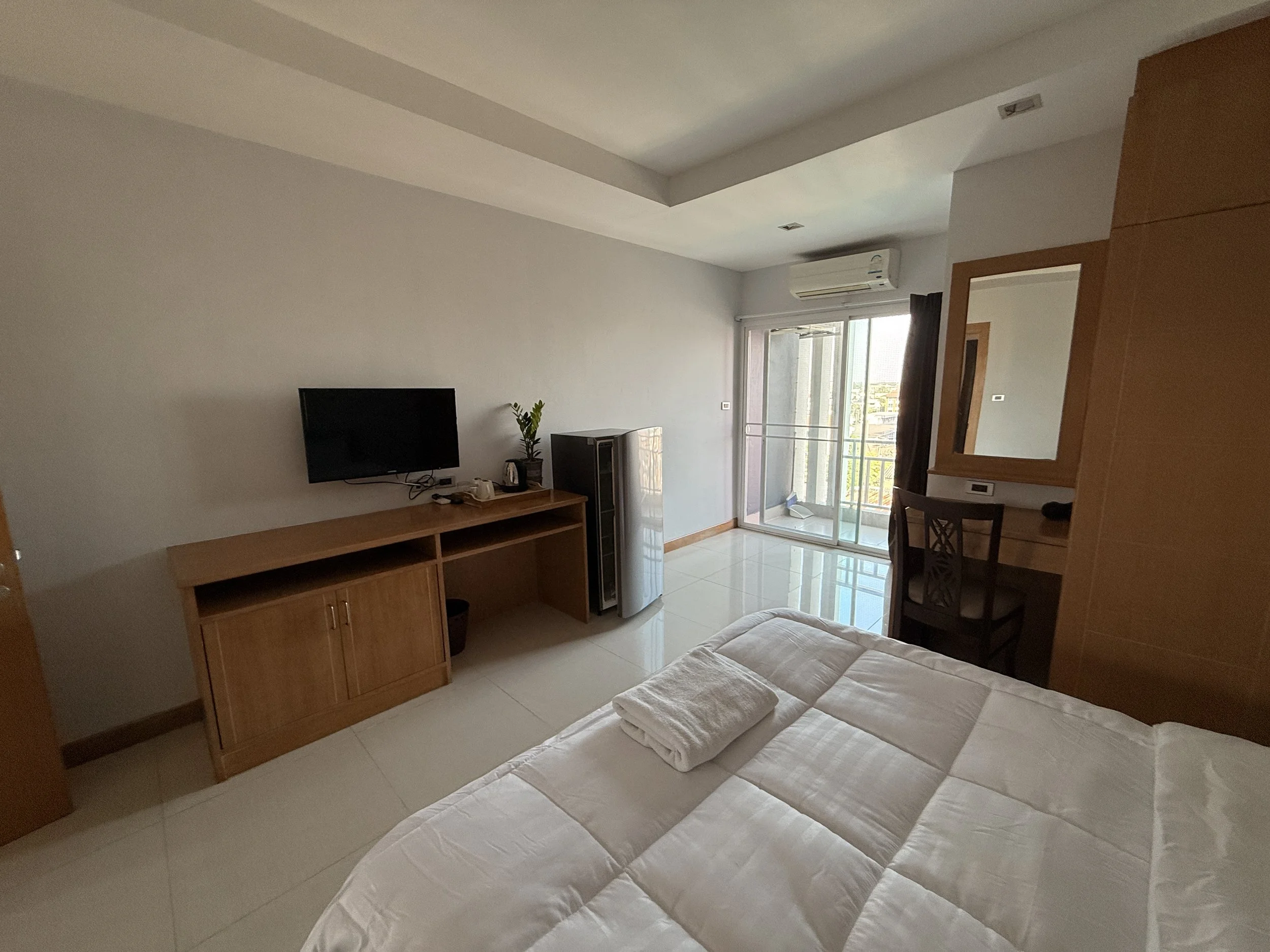 A bedroom with white tiled floors, a bed with white linens, and a towel folded on it. There is a wooden desk with a small TV, a potted plant, and a mini fridge. A sliding glass door leads to a balcony with a view of the city.