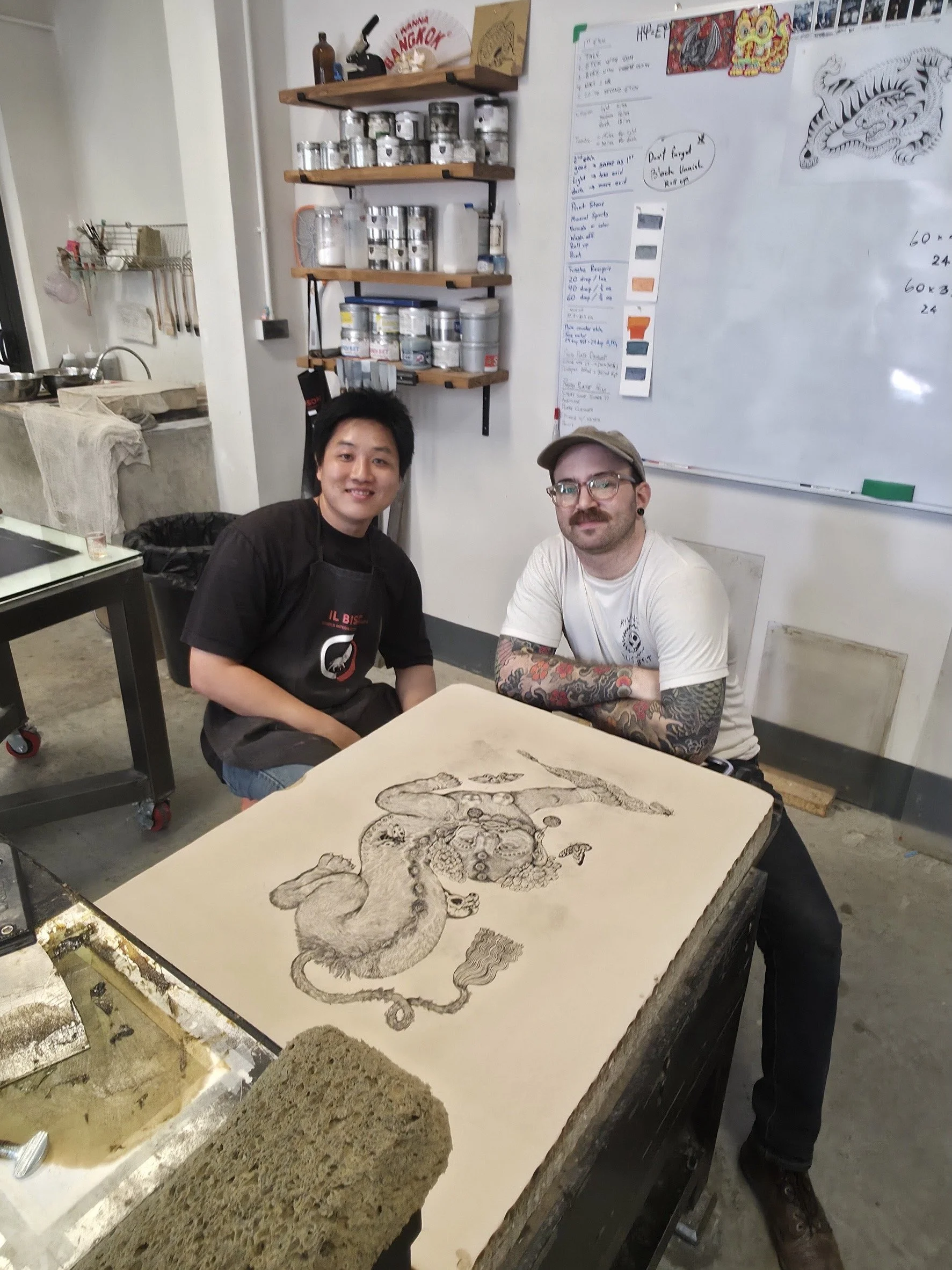 Two men sitting at a table with a detailed illustration of a lion on a piece of stone or paper in front of them. One man wears a black apron and has dark hair, while the other has glasses, tattoos on his arms, and a cap. The background features shelves with jars and containers, a whiteboard with notes and drawings, and a kitchen area.