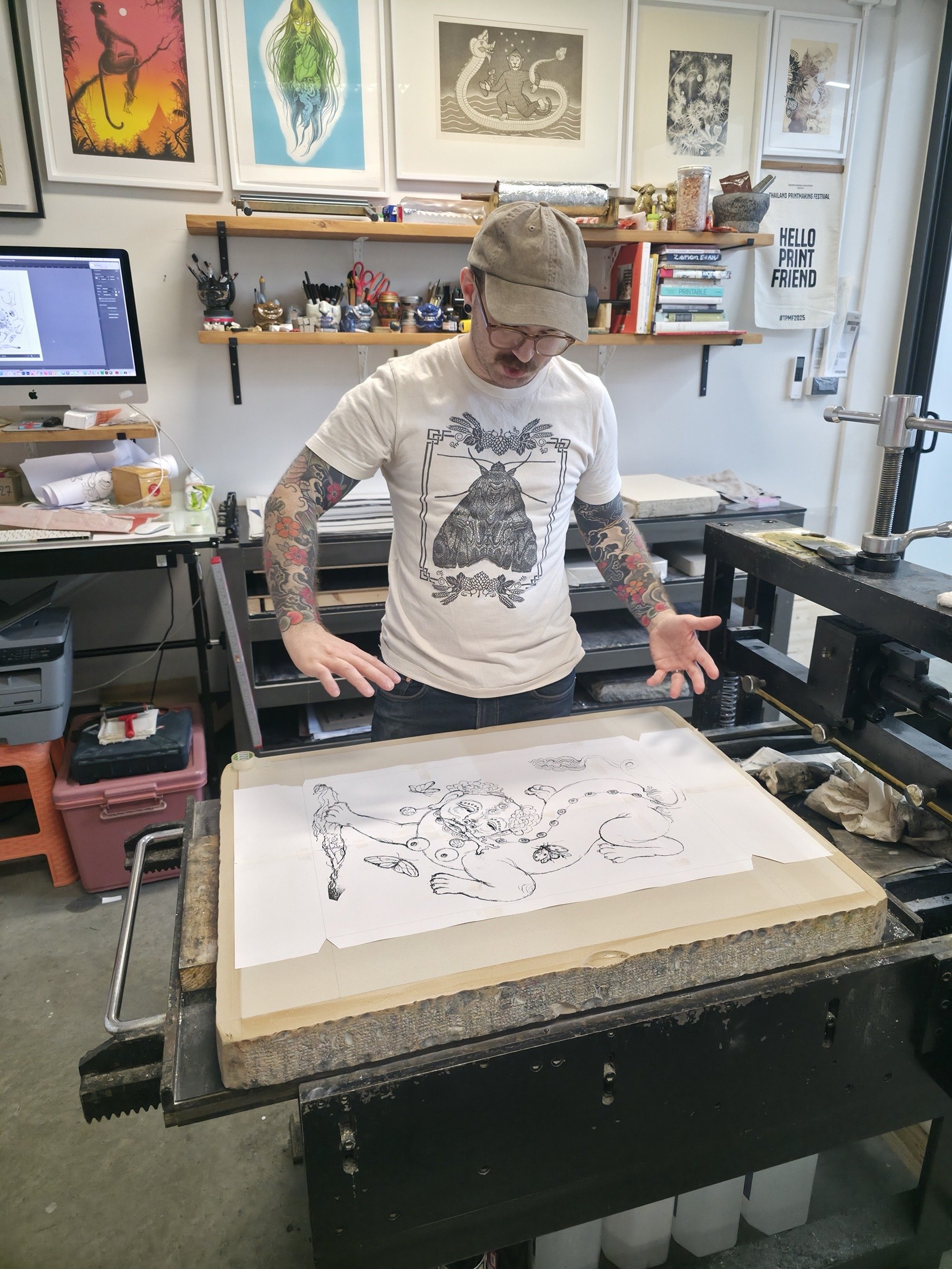 A person with tattoos on arms wearing a beige cap and glasses stands at a table with a large sheet of paper featuring a line drawing of a mythical creature. The background shows framed artwork on the wall, shelves with art supplies, and a computer on the left.