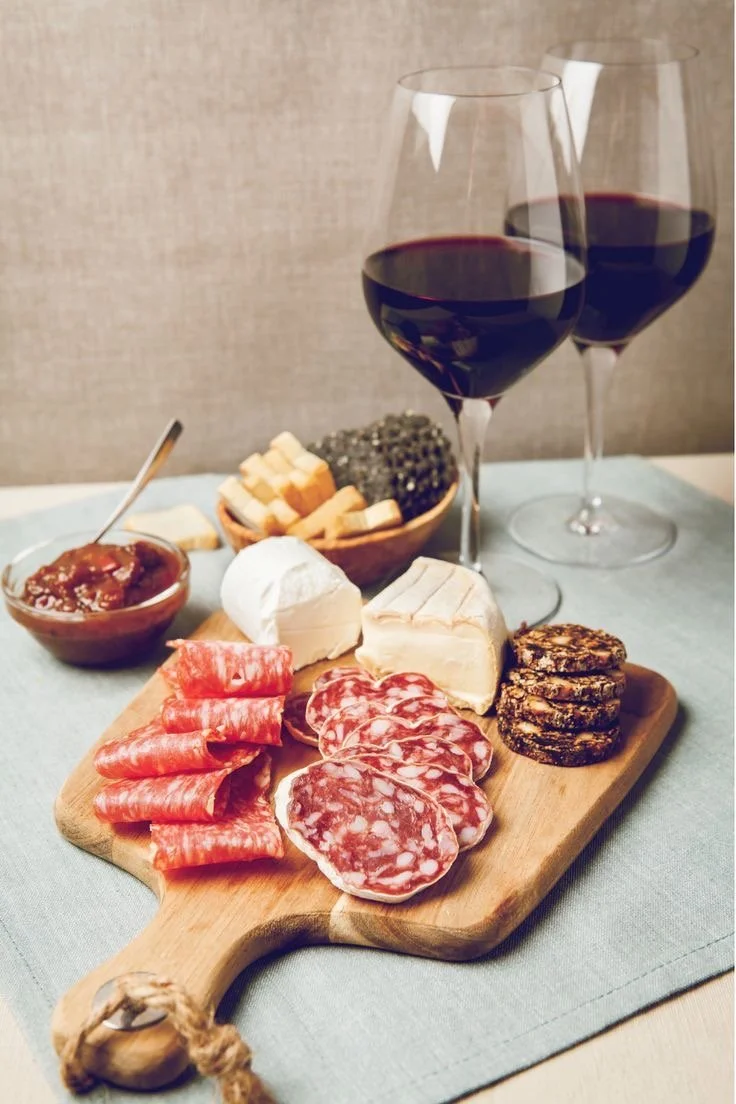 Charcuterie board with various cheeses, cured meats, and crackers, two glasses of red wine, and a bowl of jam on a table.