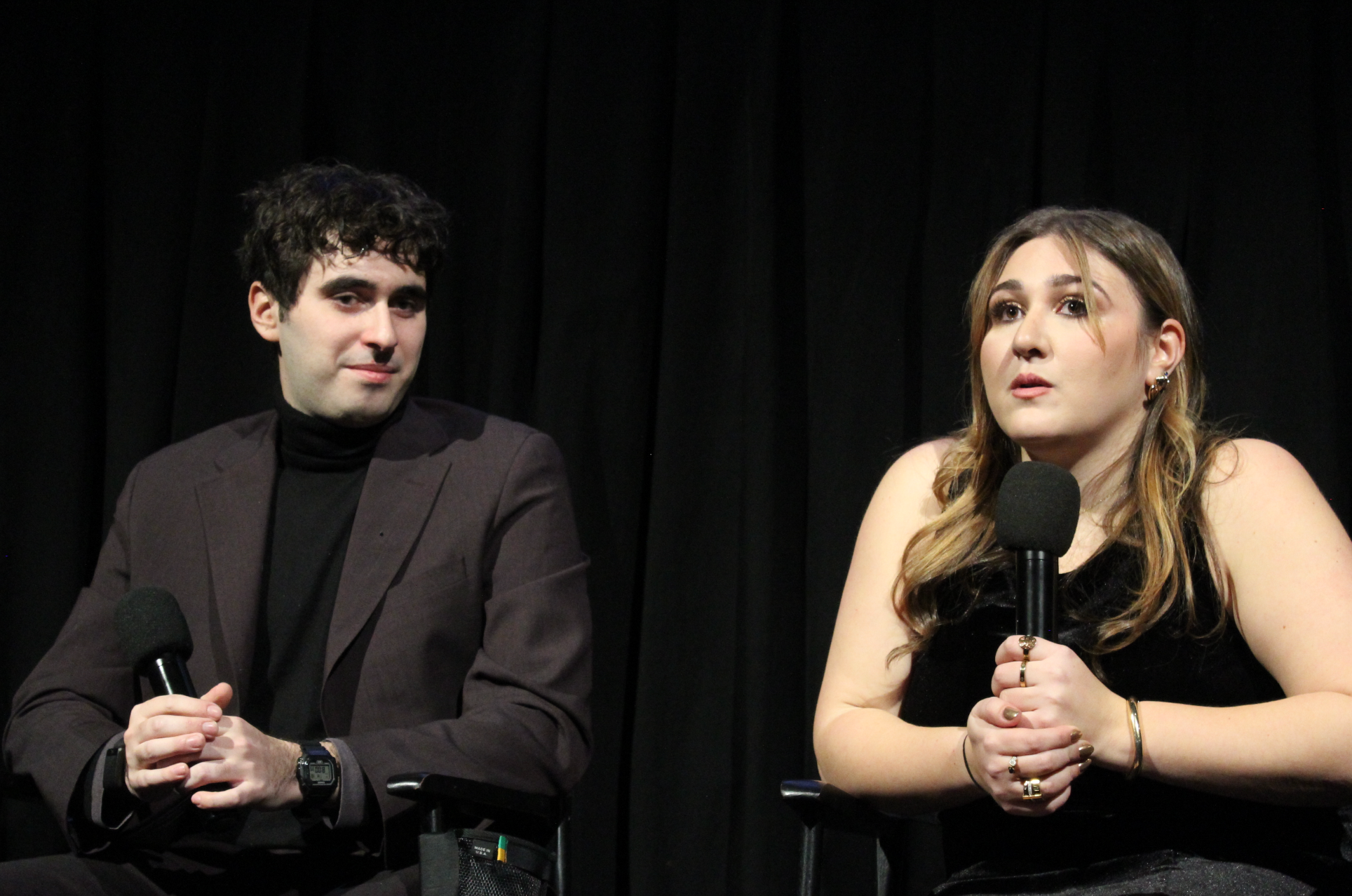 Two individuals sitting on stage, each holding a microphone. The man on the left has dark curly hair, wearing a suit and black turtleneck. The woman on the right has long light brown hair, wearing a black dress and multiple rings and bracelets. They are in front of a black curtain backdrop.