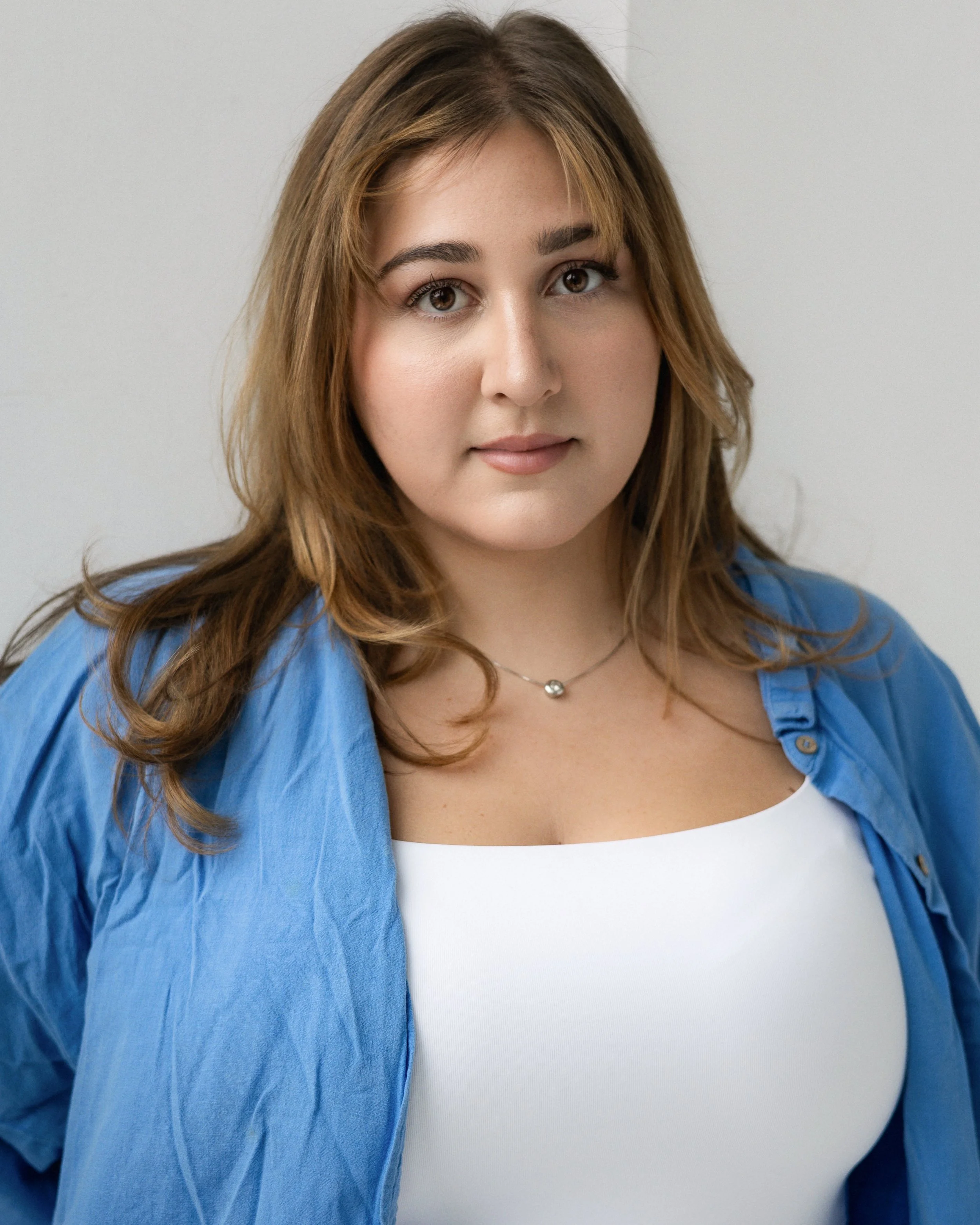 Close-up of a woman with brown hair, wearing a white top and a blue shirt, looking into the camera with a neutral expression.