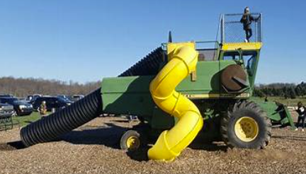 A large, colorful machine with a yellow spiral slide, green body, black hose, and large tires, set up in an outdoor area with other vehicles in the background.