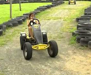 Child riding a small go-kart on a dirt path surrounded by stacks of tires at a go-kart track.