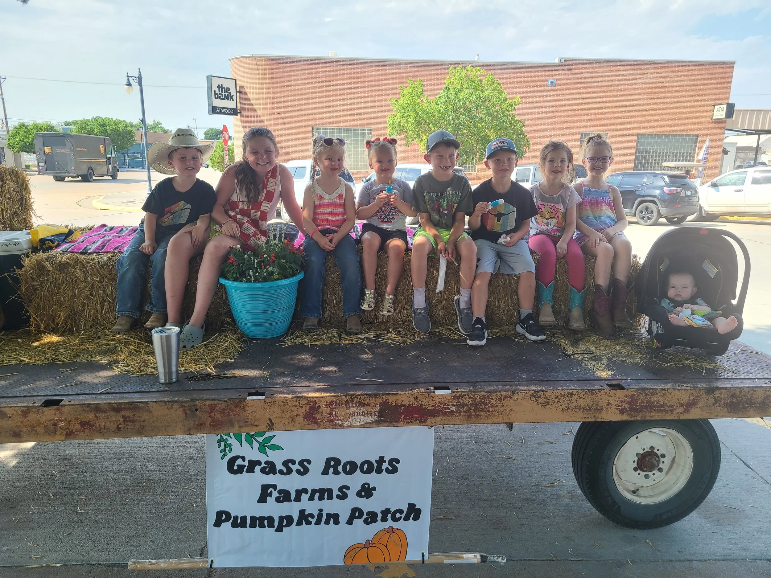 Children sitting on hay bales at a pumpkin patch with a sign that reads "Grass Roots Farms & Pumpkin Patch" in front of a brick building with trees and parked cars in the background.
