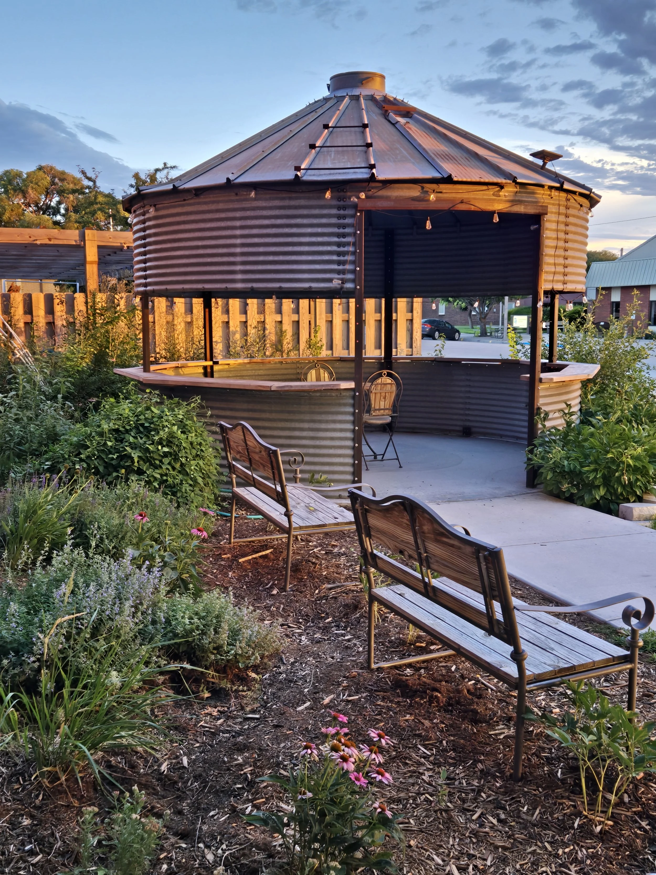 A circular outdoor seating area with chairs, surrounded by plants and flowers, under a wooden and metal covered structure with outdoor string lights at dusk.