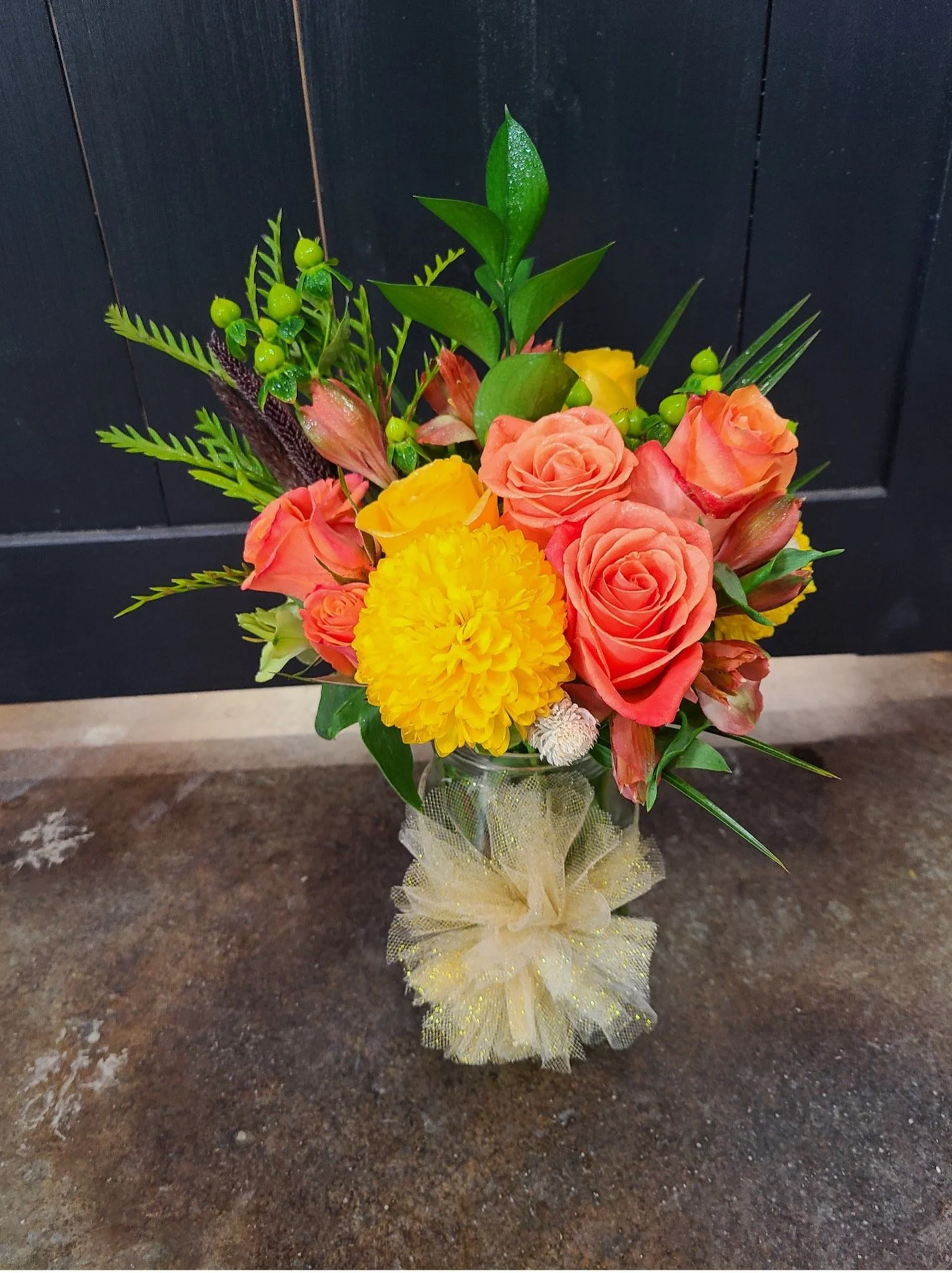 A bouquet of mixed flowers in a glass vase with yellow, orange, and pink roses, yellow marigold, and green foliage, wrapped with a gold mesh ribbon, placed on a brown surface against a black background.