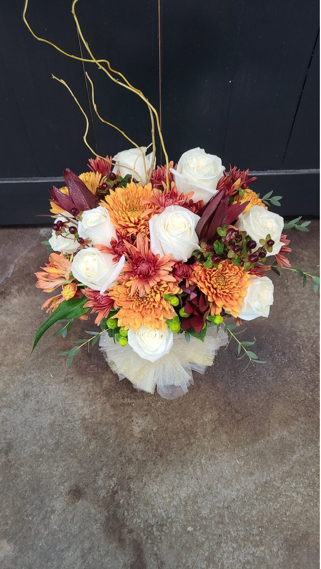 A bouquet of flowers with white roses, orange chrysanthemums, deep red leaves, green berries, and decorative yellow twigs, wrapped in a mesh ribbon.