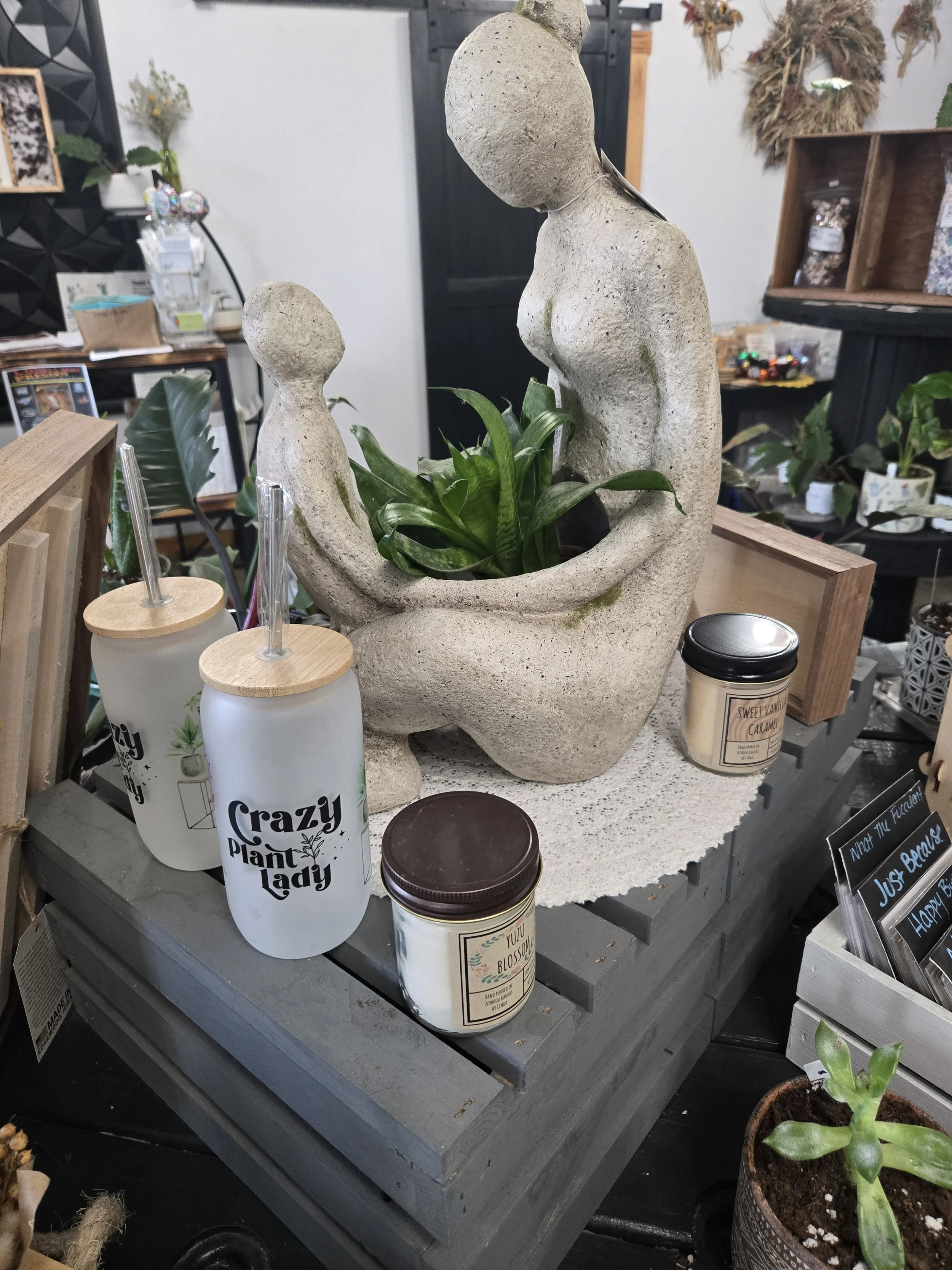A decorative stone sculpture of a mother holding a baby, with a green plant in the middle, displayed on a table in a store surrounded by other plants and jars.