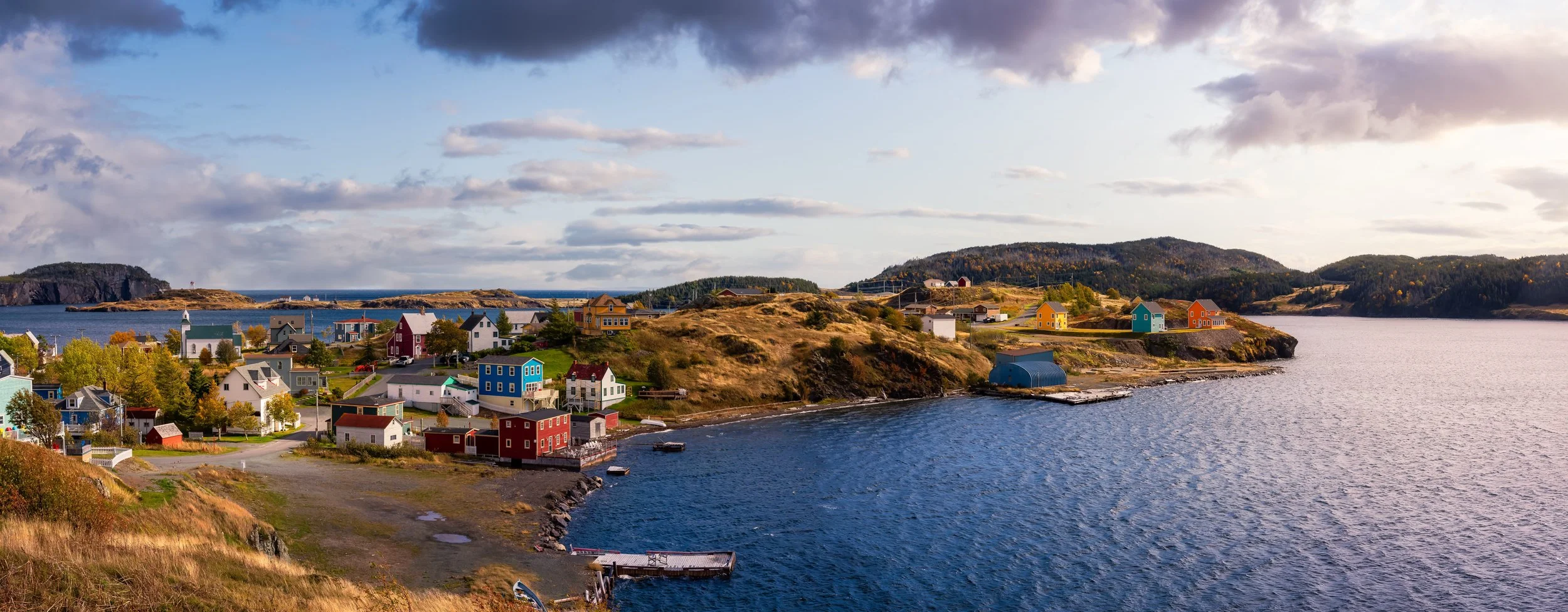 Colorful houses on a hill by the water in a coastal village, with hills and cloudy sky in the background.
