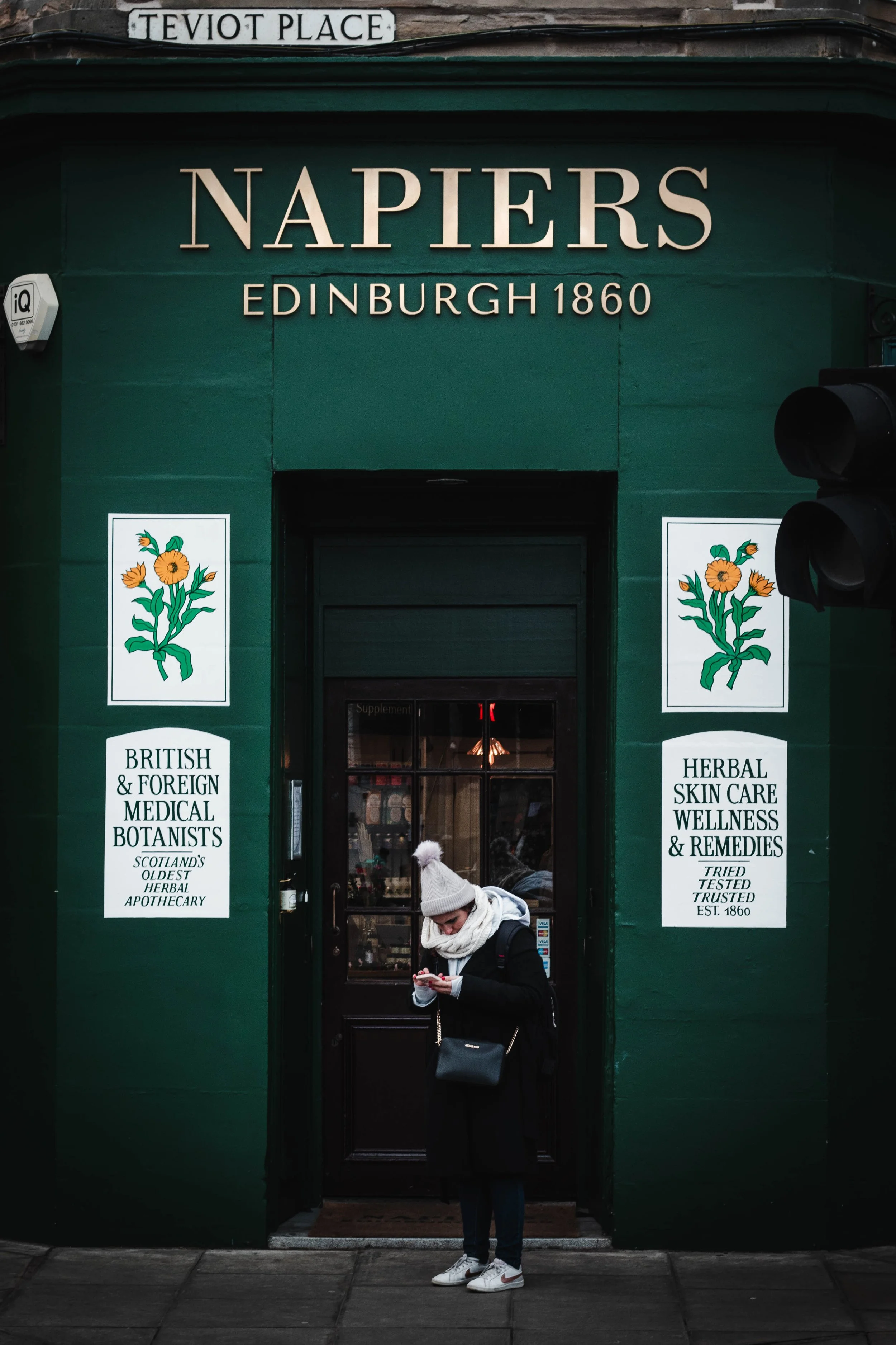 Person in winter clothing standing in front of Napiers herbal medicine shop in Edinburgh, established in 1860, with green exterior and floral signs.
