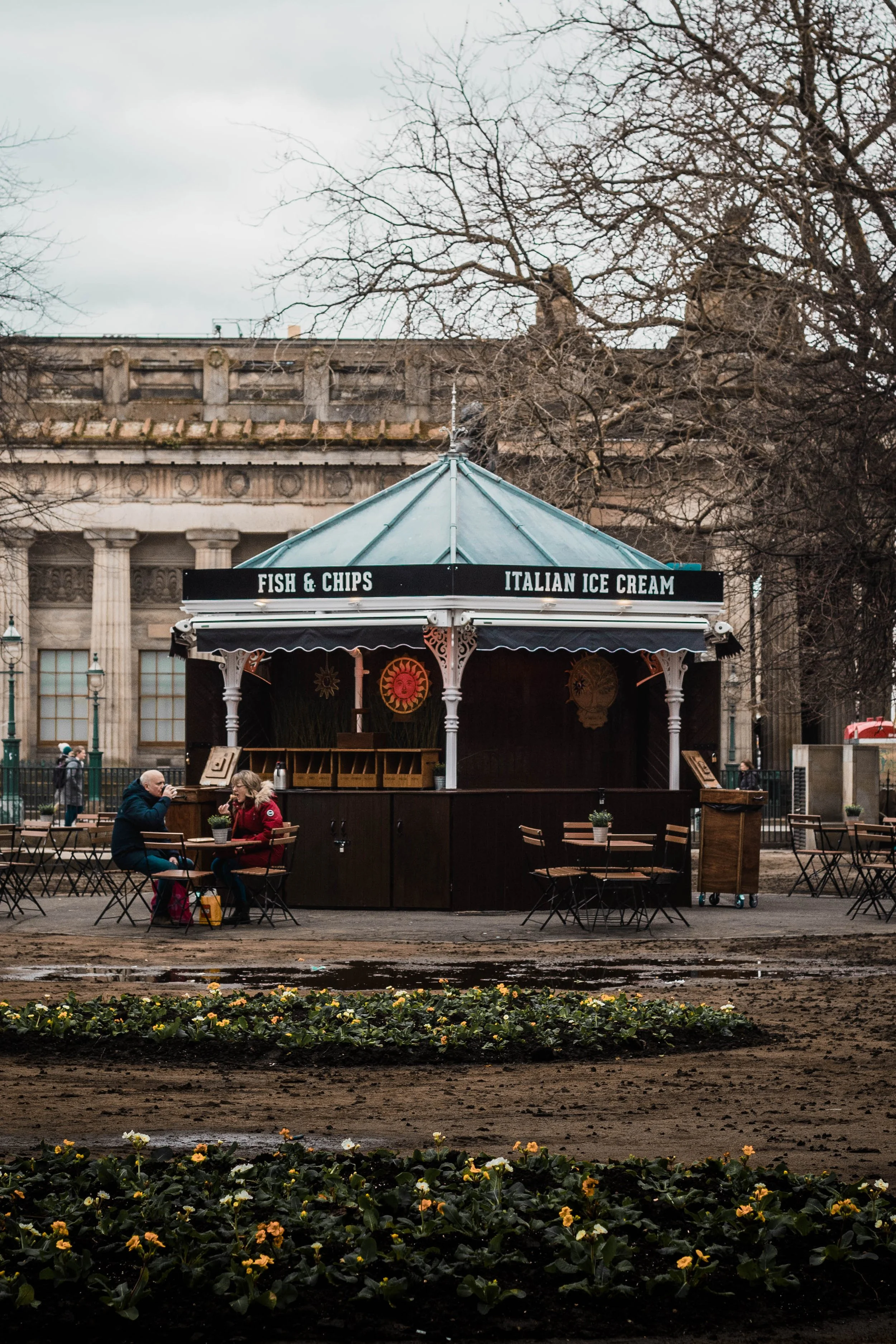 A small outdoor stand with a sign reading 'Fish & Chips' and 'Italian Ice Cream' against a background of leafless trees and a historic building. Two people are sitting at a table near the stand, talking.