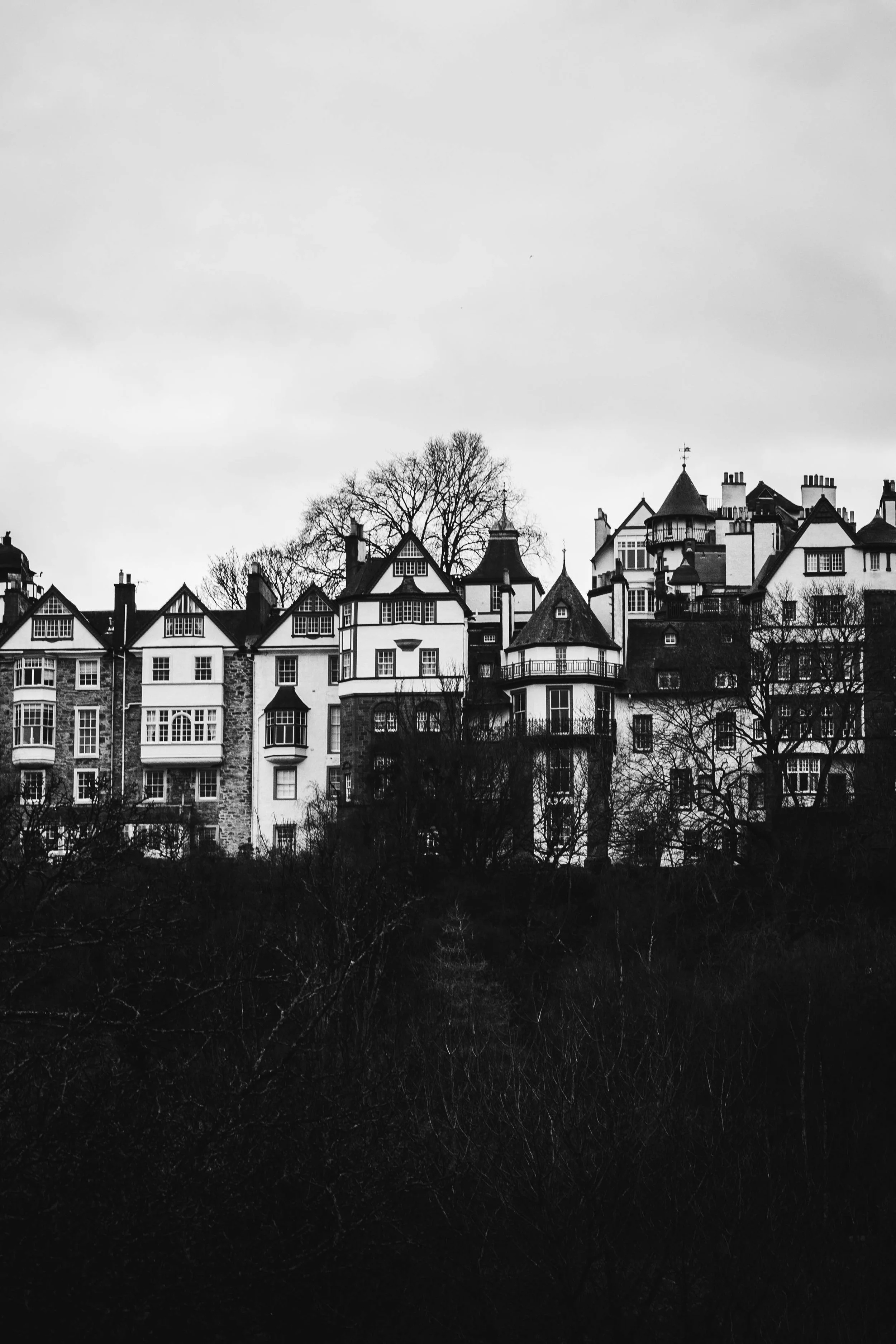 Black and white photo of a hillside with a row of old, multi-story houses with steep roofs and chimneys, surrounded by leafless trees.