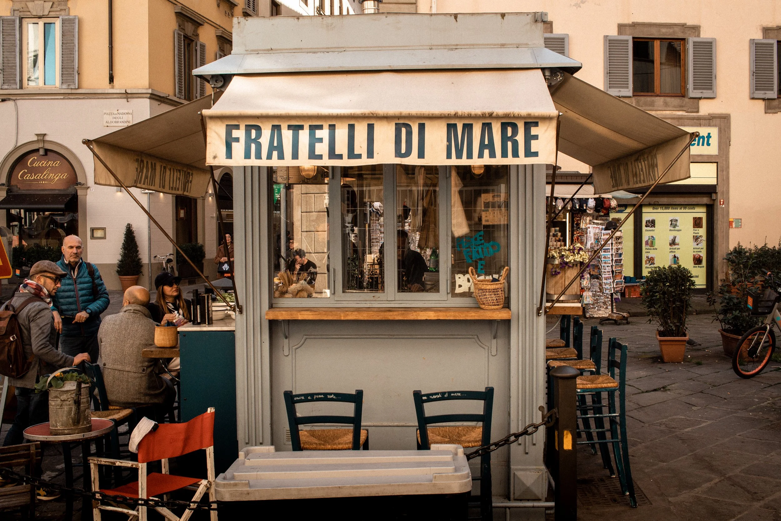 Outdoor kiosk with a sign reading 'FRATELLI DI MARE' in a European city square surrounded by shops, with people sitting and standing around.