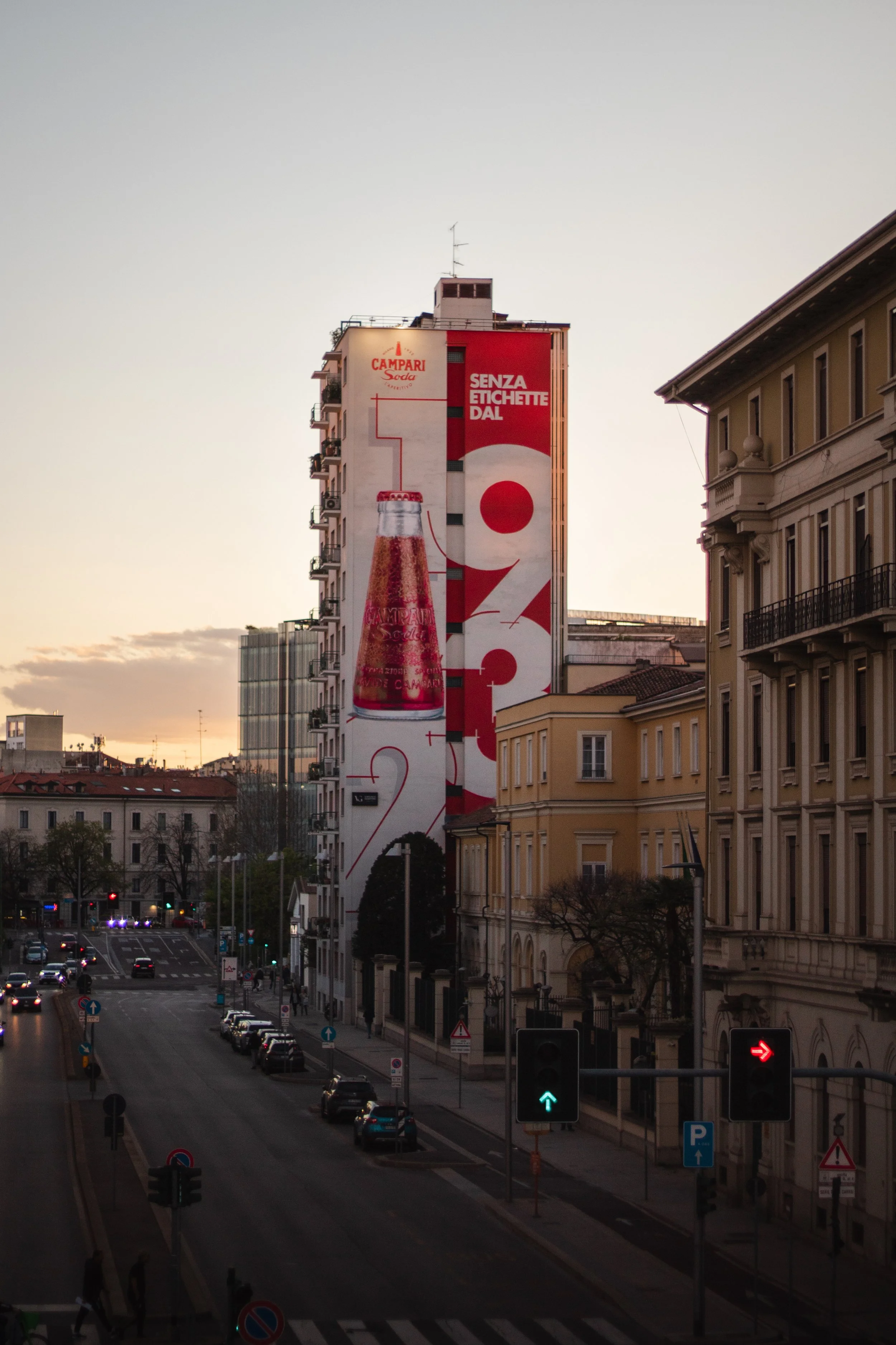 Tall building with a large vertical advertisement for Campari soda featuring a red and white color scheme, a perfume bottle, and Italian text, with surrounding city streets and buildings at dusk.