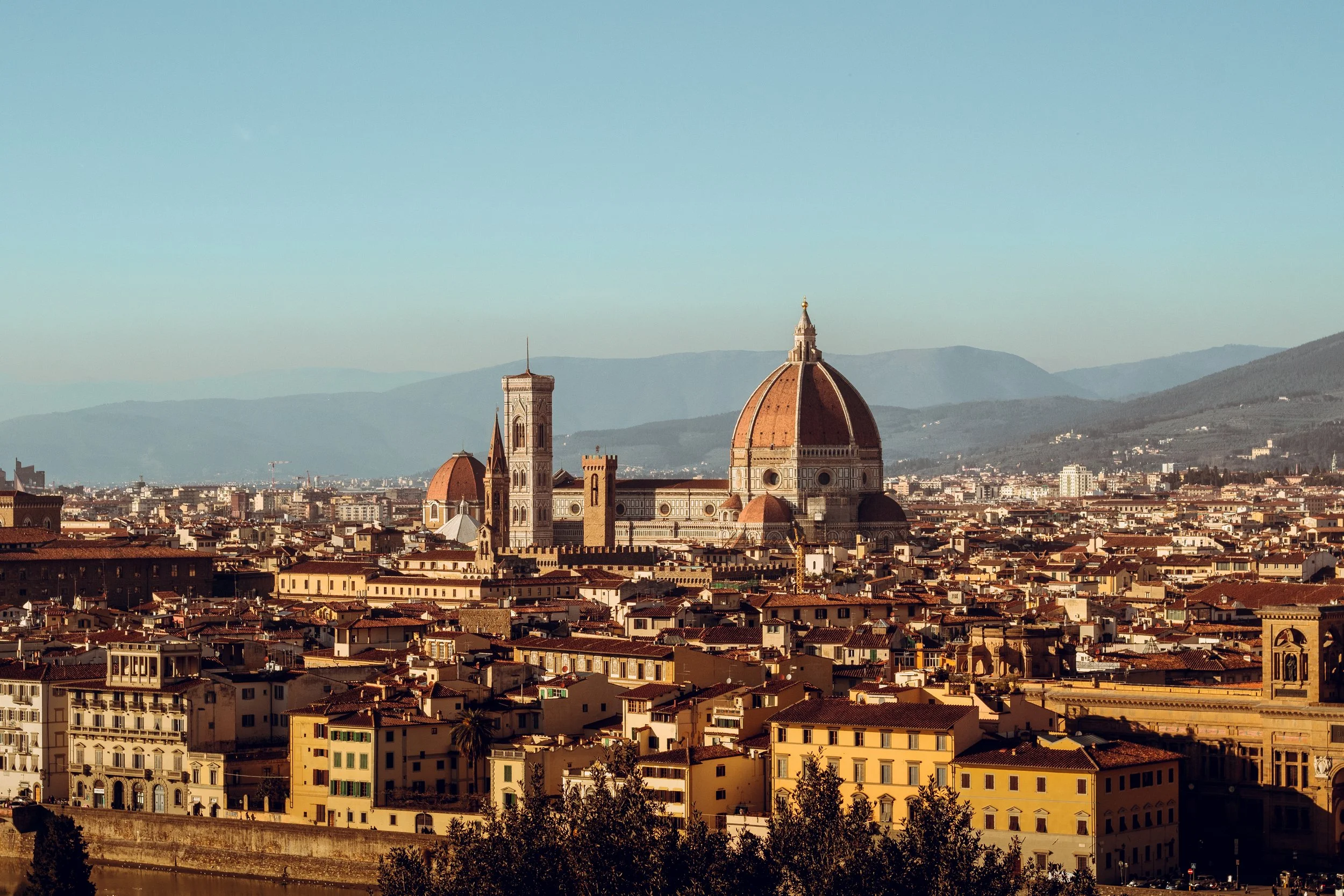 View of Florence, Italy, featuring the Florence Cathedral with its large dome and surrounding historic buildings, with mountains in the background.