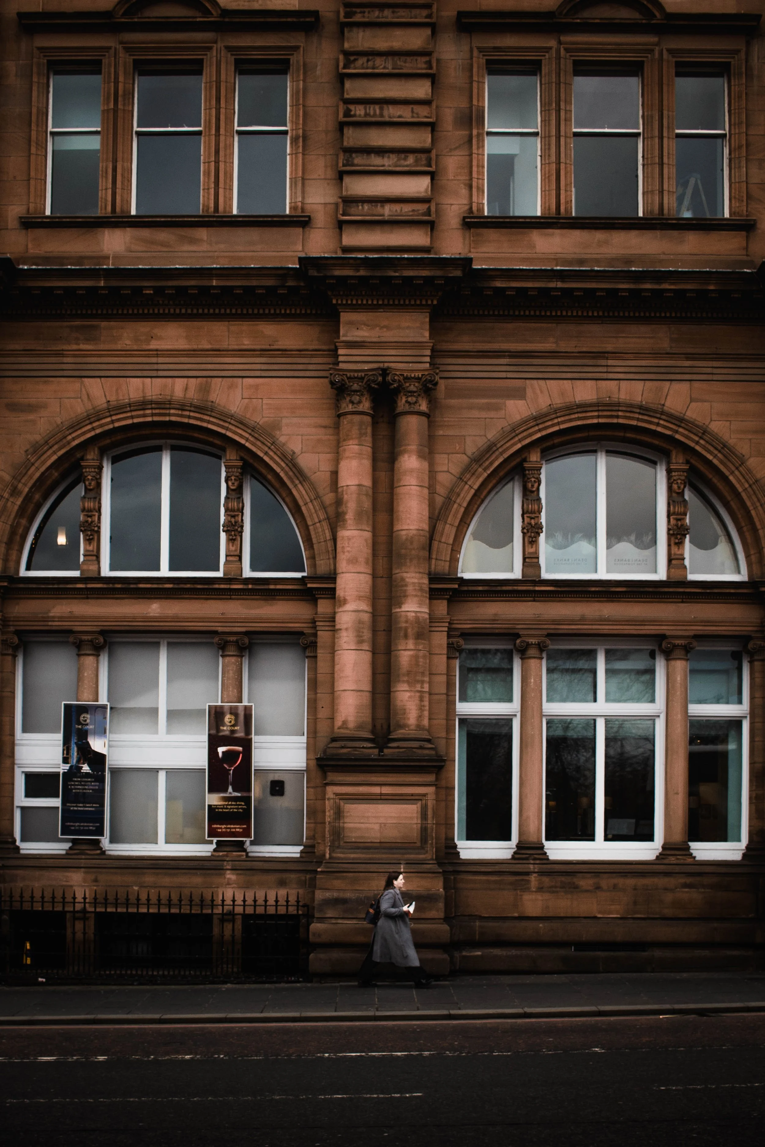 A person walking past a large ornate building made of red-brown stone, with big arched windows and decorative columns, on a city street.