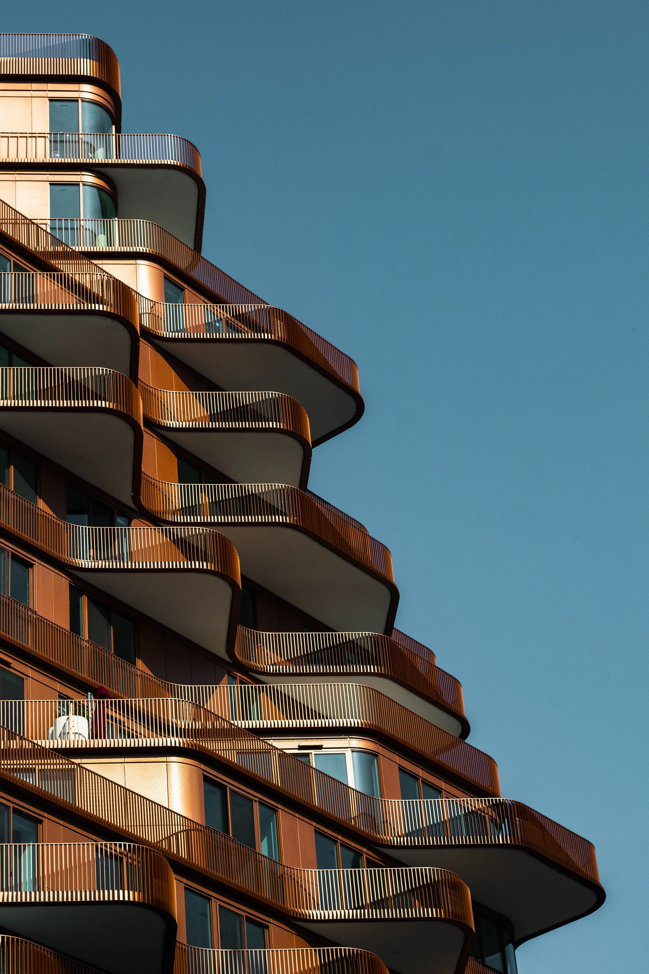 A modern high-rise building with curved balconies featuring metal railings, set against a clear blue sky.