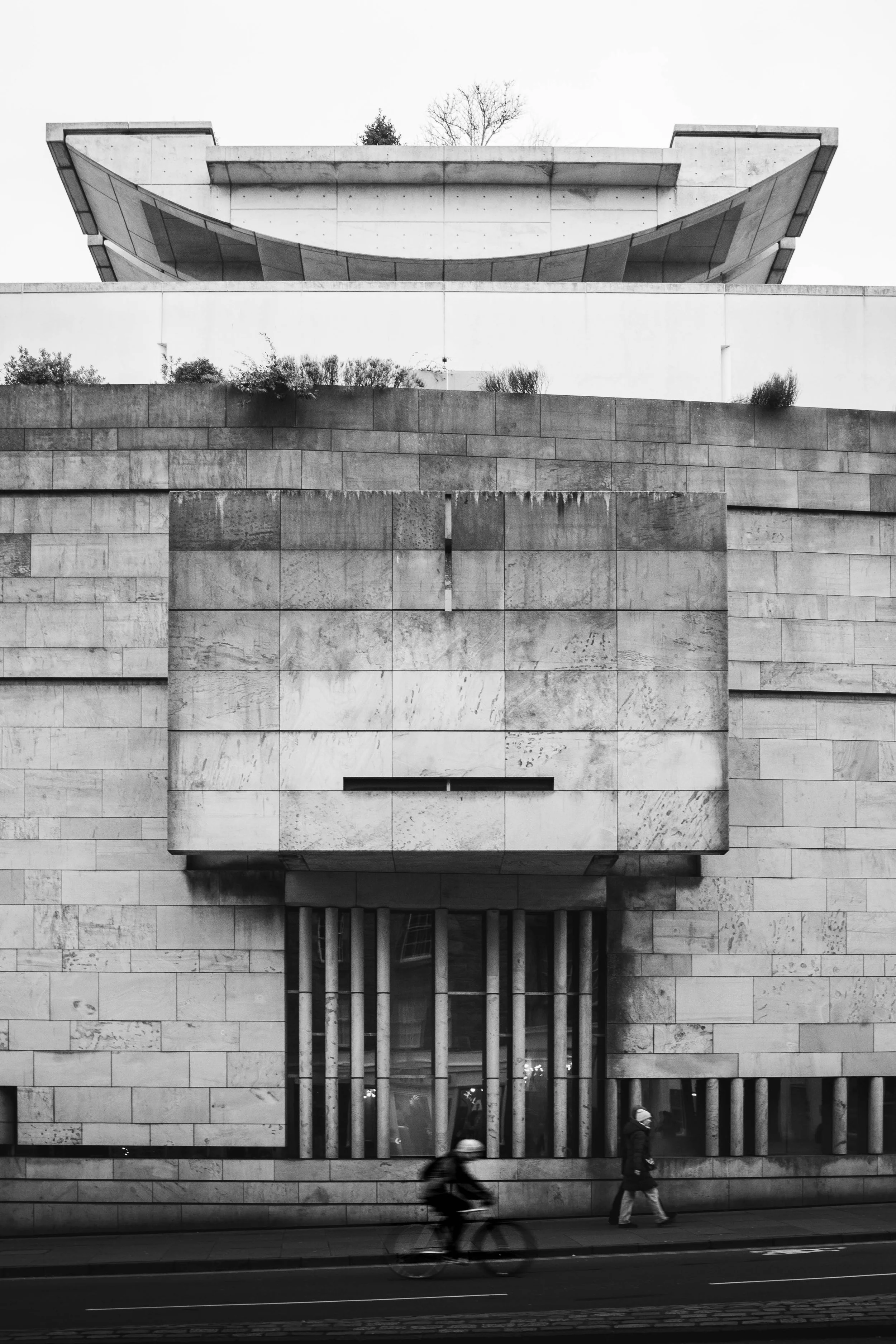 Black and white photo of a modern building with geometric architecture, featuring large rectangular stone blocks, vertical metal bars, and a person and cyclist passing by on the street in the foreground.